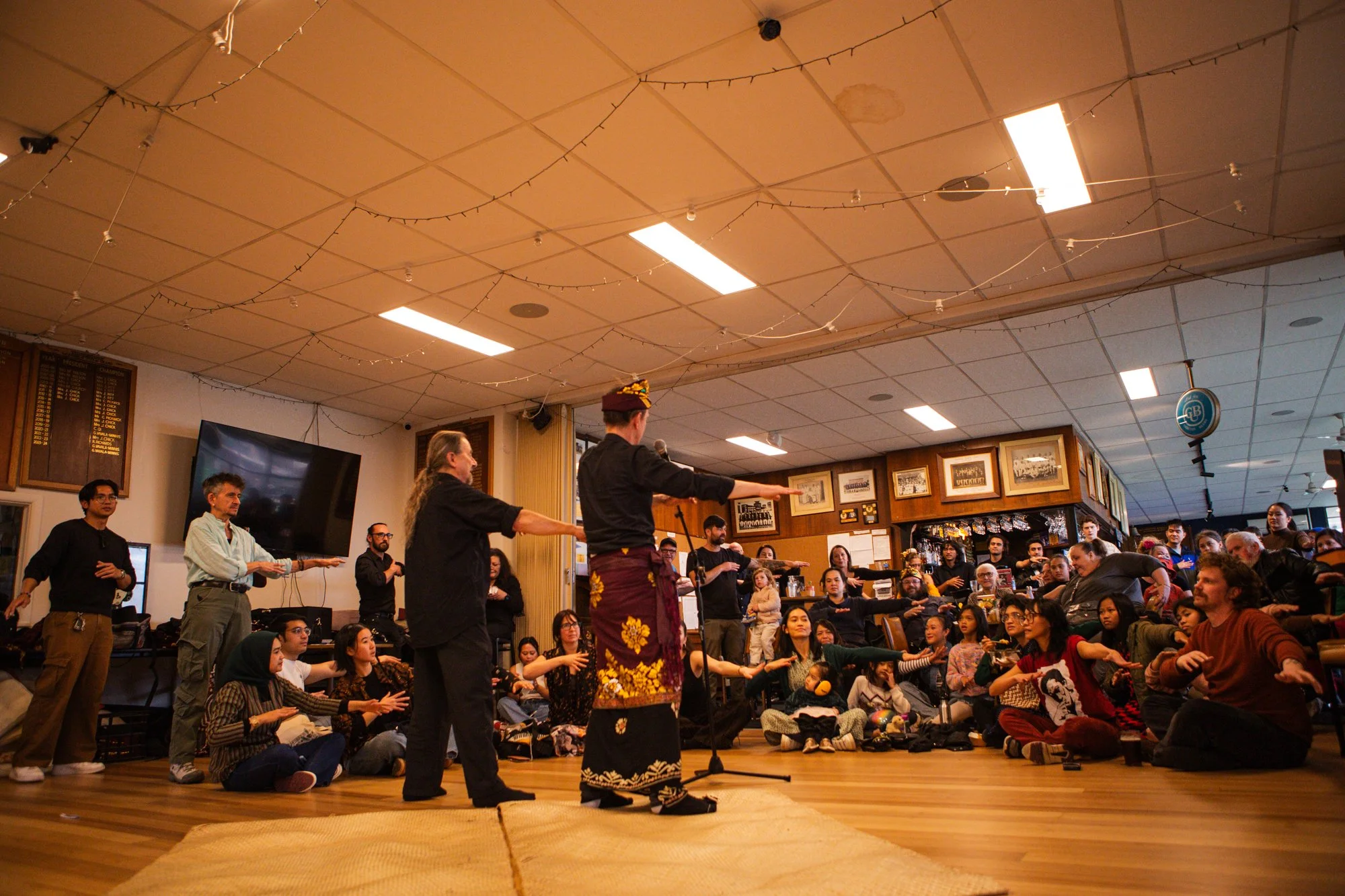 Performers in traditional attire leading a dance or gesture in front of an audience sitting on the floor and standing in a room decorated with framed pictures, string lights, and a large television screen.