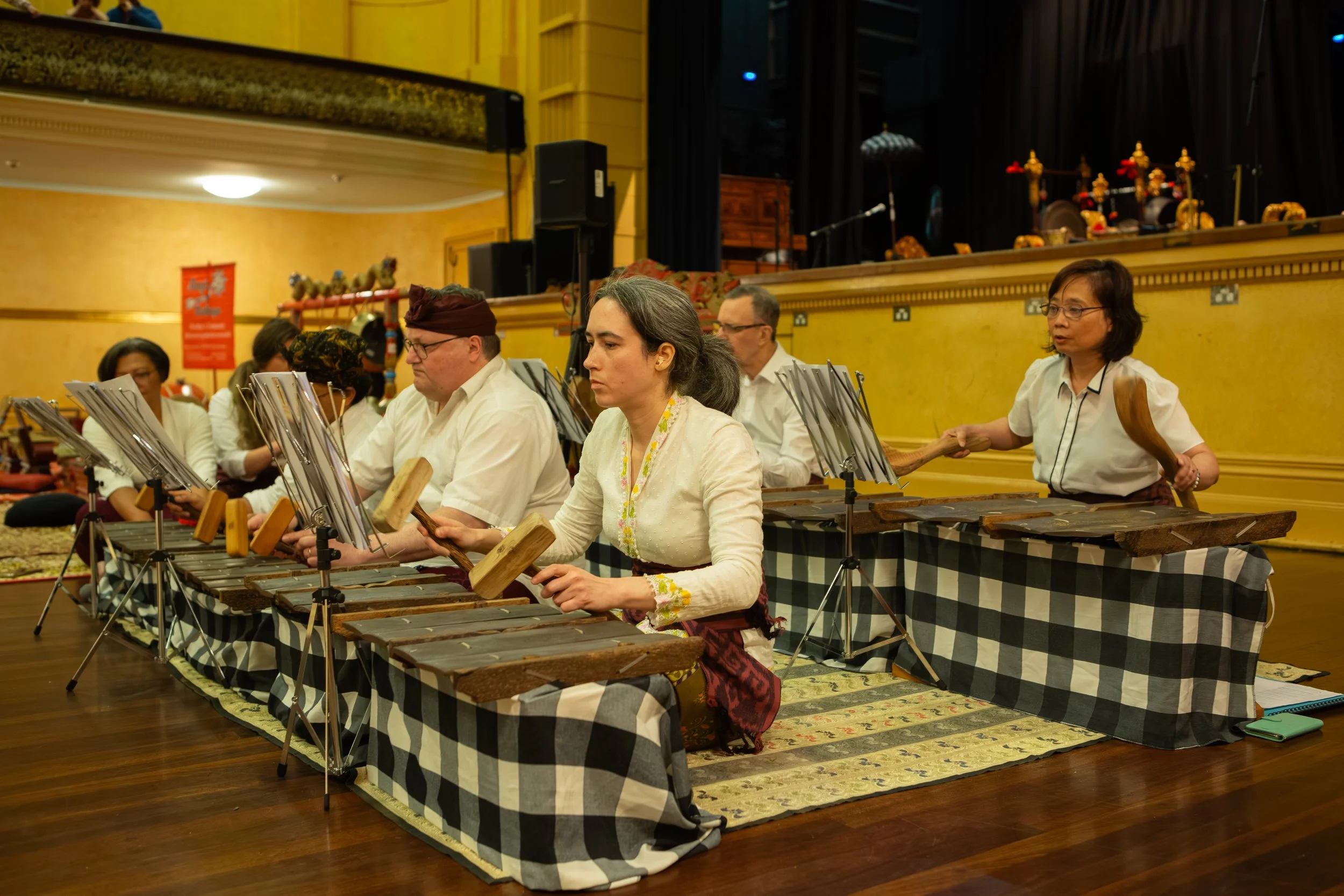 A group of musicians playing traditional Indonesian gamelan instruments, sitting on a checkered cloth-covered platform in a performance hall.