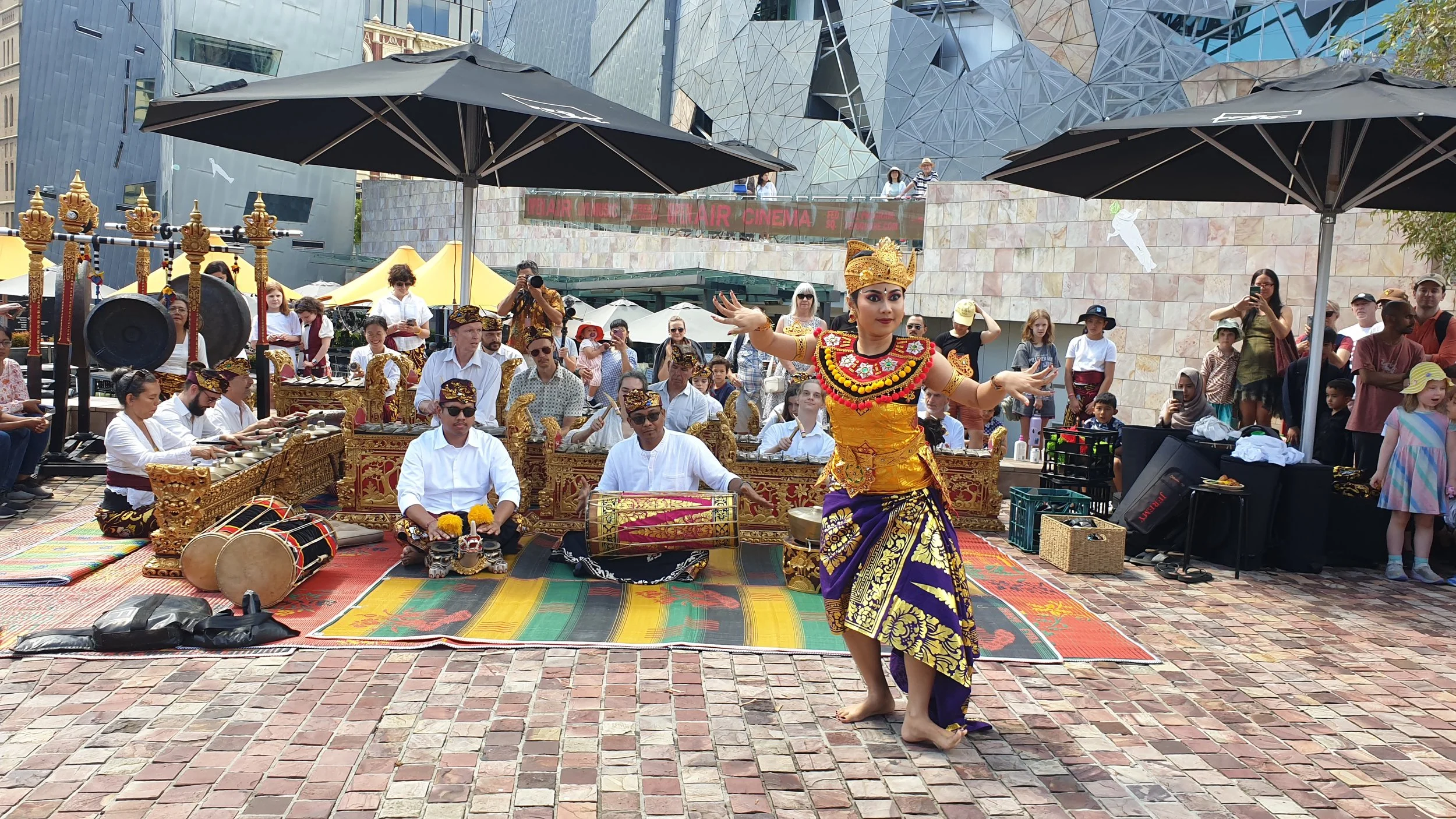 A traditional Balinese dance performance outdoors with musicians playing instruments behind a female dancer. The dancer is dressed in colorful, ornate costume with gold accents, performing on a brick-paved ground, under banners and umbrellas, surroun