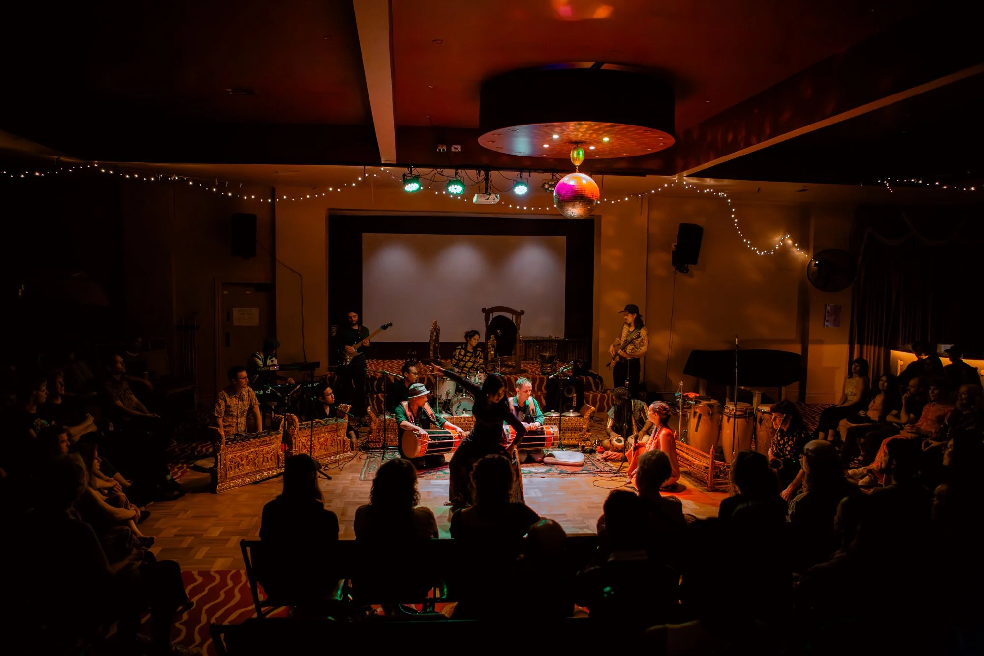 A live music performance in a dimly lit venue with audience members seated around a stage. A band with various instruments plays on stage, including drums, guitars, and percussion. The ceiling has string lights and a disco ball.