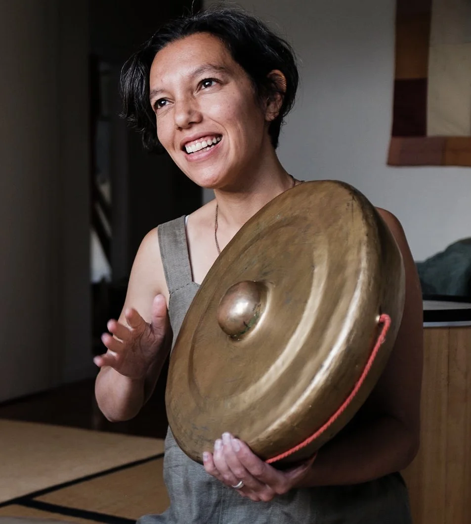 A woman with short black hair smiling and holding a large brass singing bowl indoors.