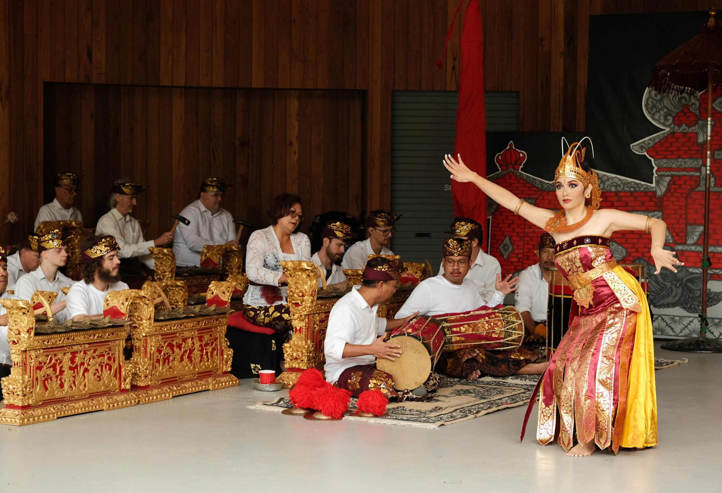 Traditional Balinese dance performance with a female dancer in ornate costume and a group of musicians playing gamelan instruments in a wooden-finished indoor setting.