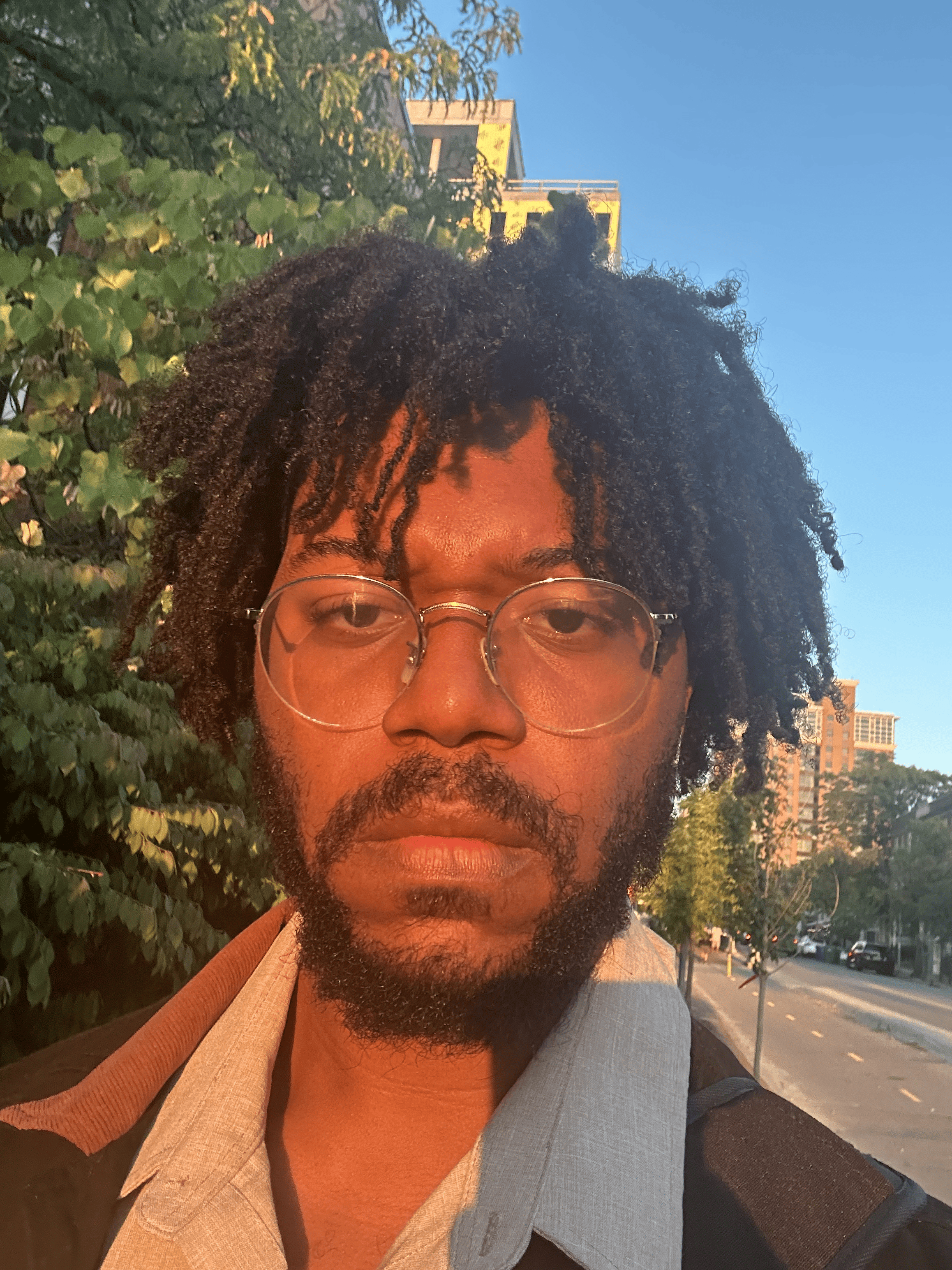 A man with glasses and a beard, wearing a light-colored shirt and carrying a backpack, standing outdoors during sunset with trees and buildings in the background.