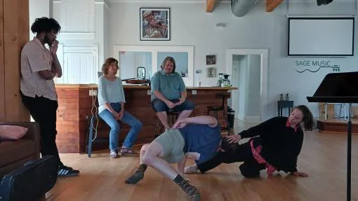 Four women in a living room watching one woman perform a wrestling move on another woman on the floor.