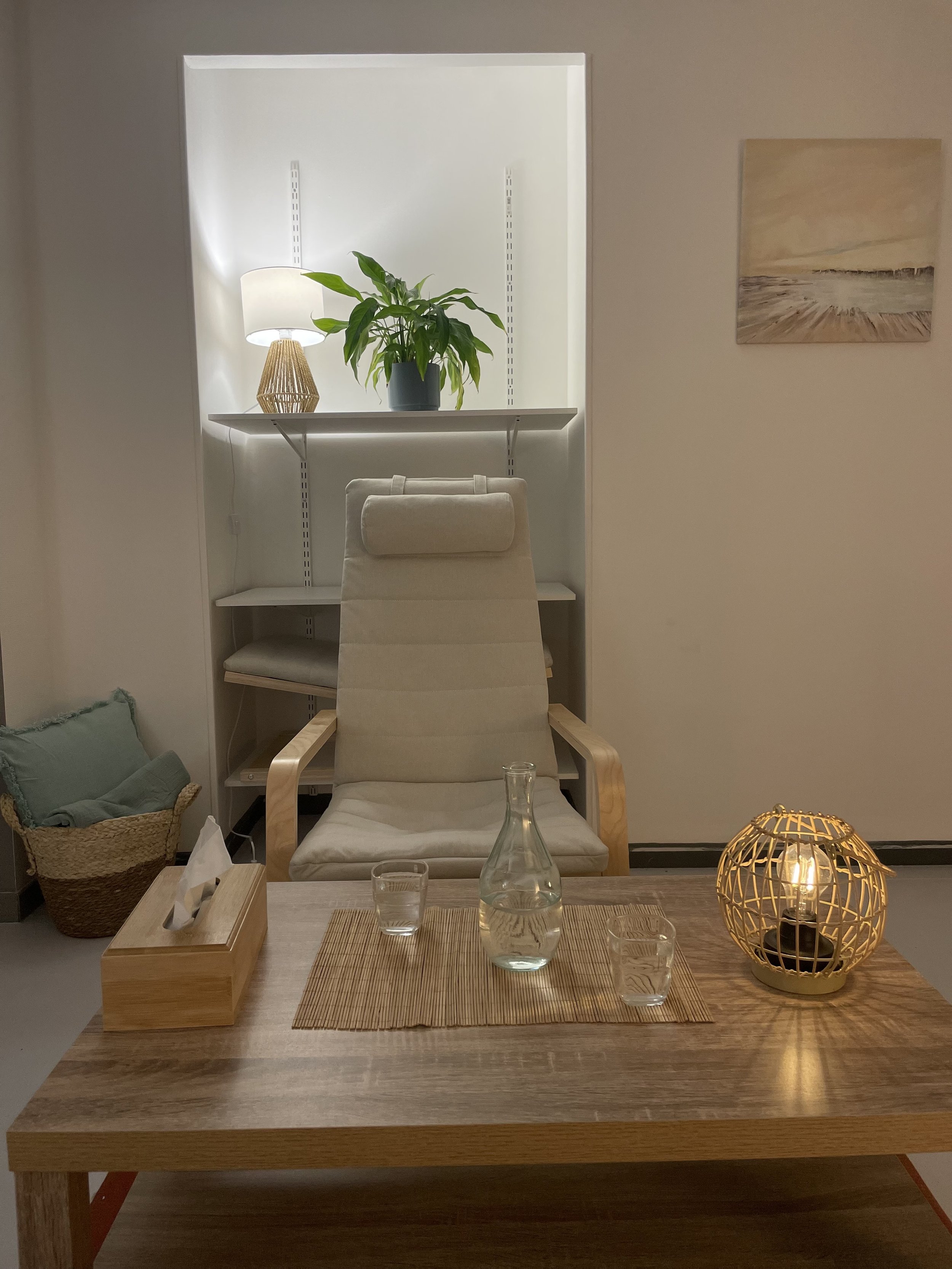 Un bureau avec une chaise beige, un plateau en bois, une carafe en verre, deux verres, une boîte en bois pour mouchoirs, une lampe en cage dorée, une peinture abstraite au mur et une étagère avec une plante verte et une lampe