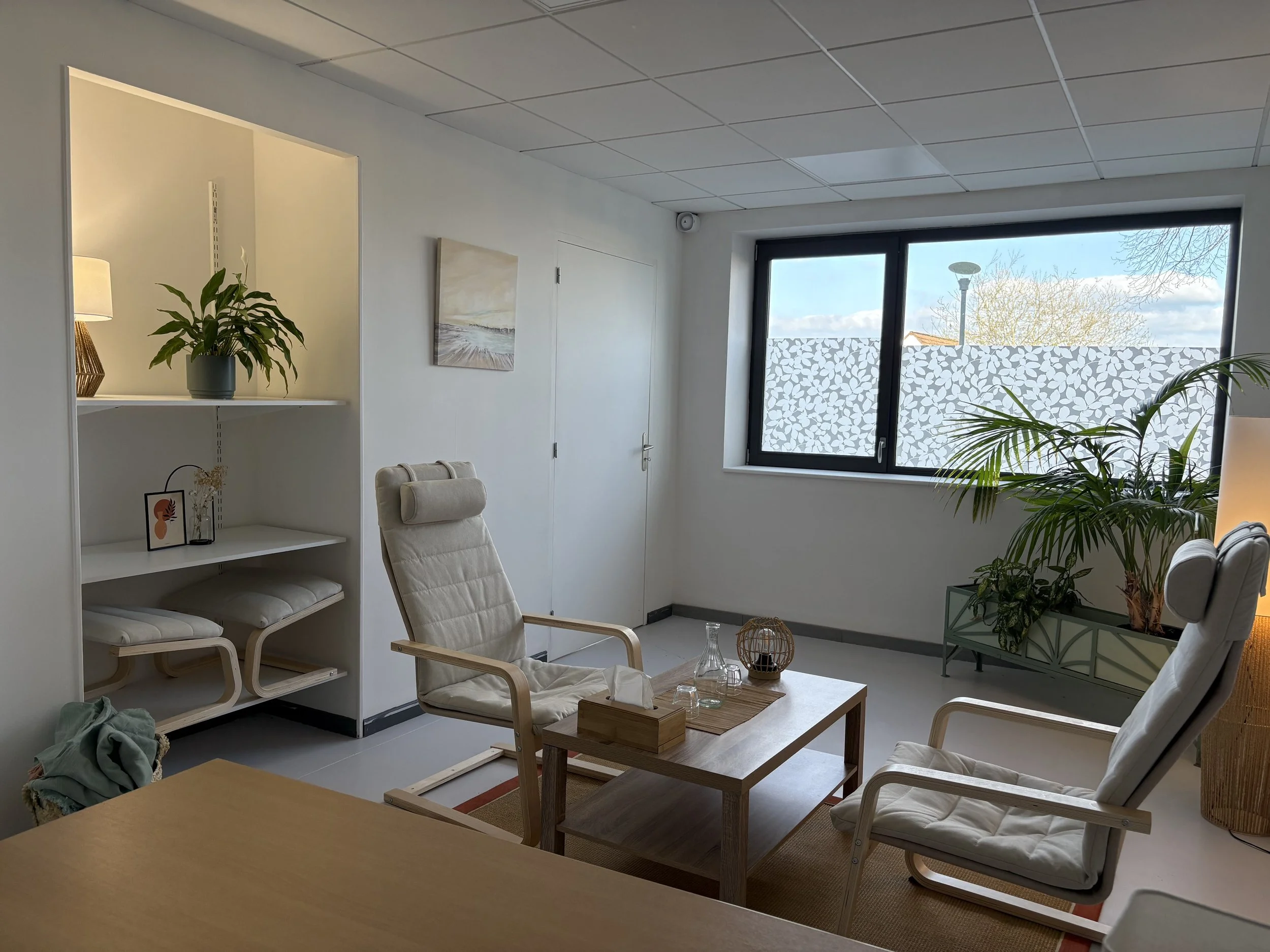 Salon lumineux avec deux fauteuils en bois et tissu, une table en bois, des plantes vertes, une grande fenêtre avec une décoration en motif de feuilles, et une étagère blanche avec des décorations.