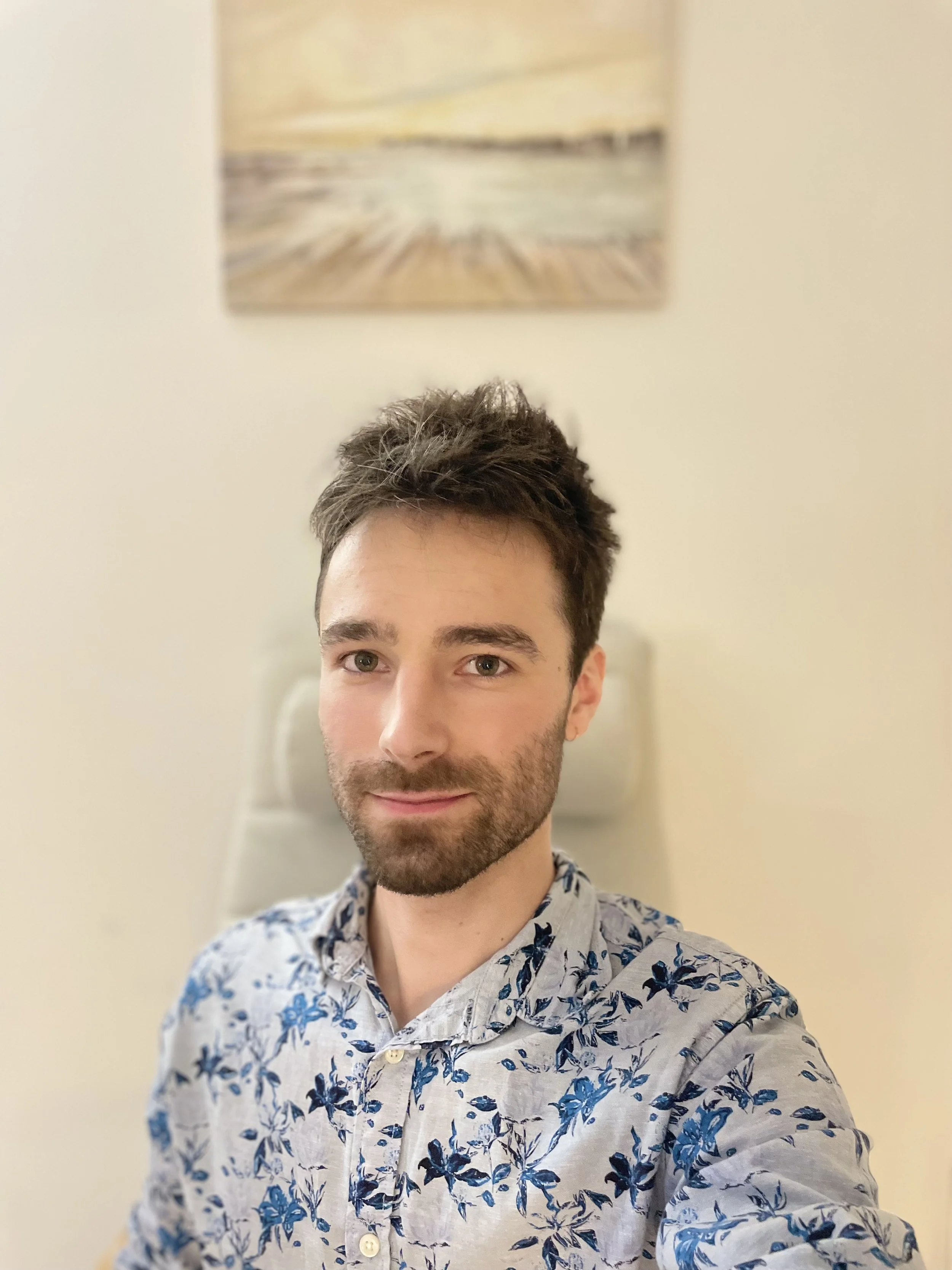 Un homme avec une barbe, portant une chemise à fleurs, posant pour une photo en intérieur, avec un tableau abstrait en arrière-plan.