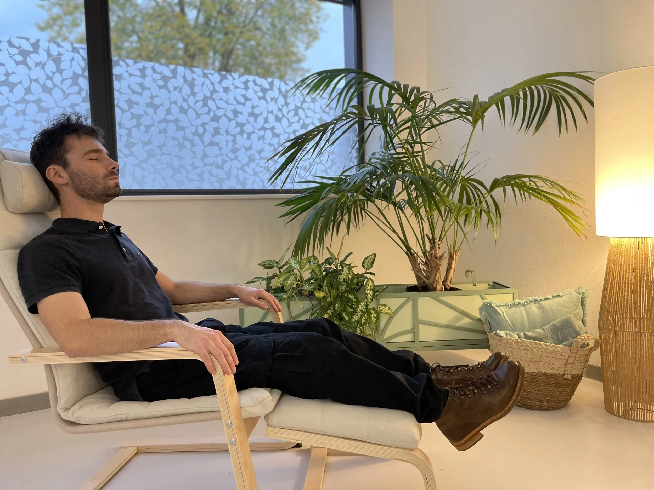 Un homme assis sur une chaise confortable avec les yeux fermés, dans une pièce calme et bien éclairée, entouré de plantes vertes et d'une lampe de lecture