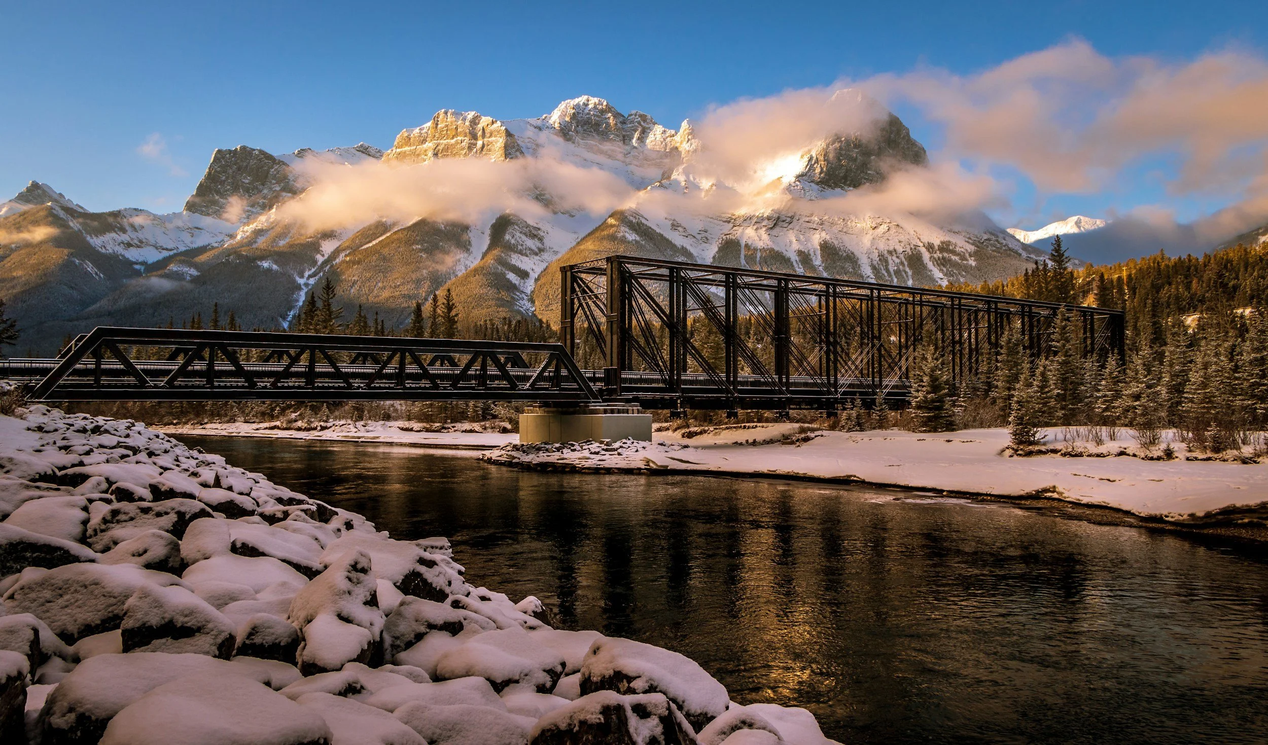 Snow-covered rocks by a river with a black metal bridge and snow-capped mountains in the background, trees along the riverbank at sunset.