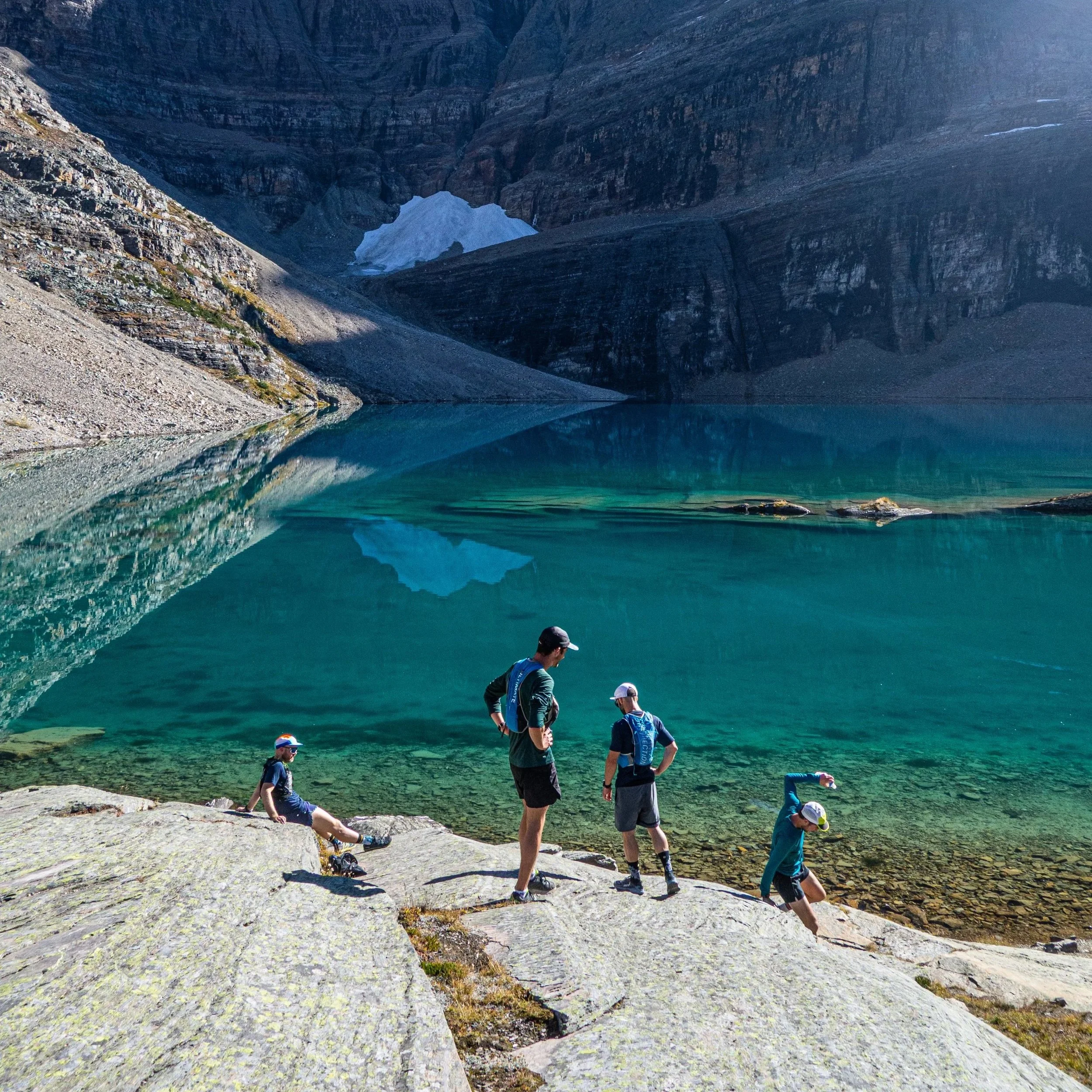 Four children and two adults standing and sitting on rocks at the edge of a clear mountain lake, with mountains and glaciers reflected in the water.