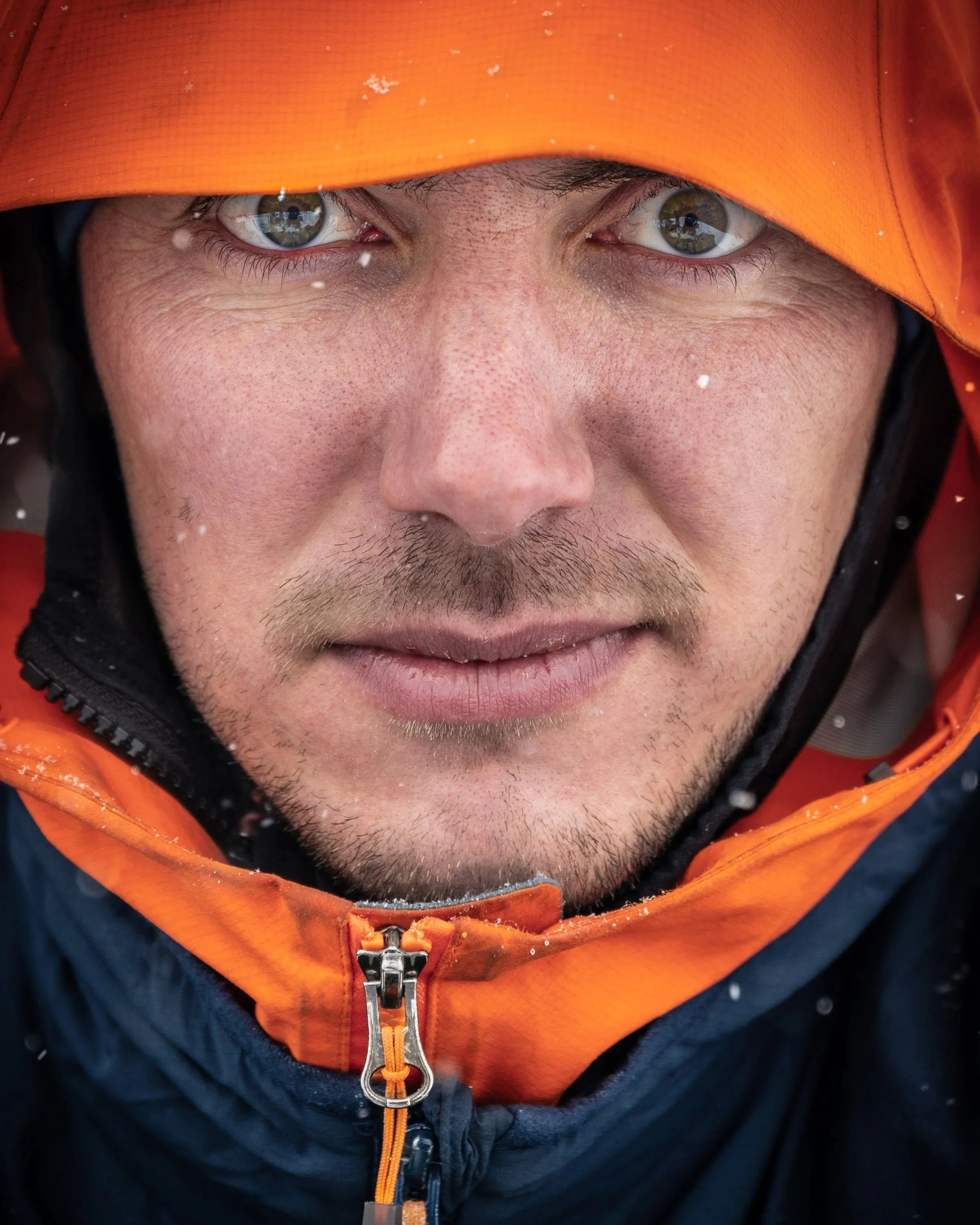 Close-up of a man in Arc'teryx outdoor gear, wearing an orange hooded jacket, with green eyes and facial hair, looking directly at the camera with a slight smile.