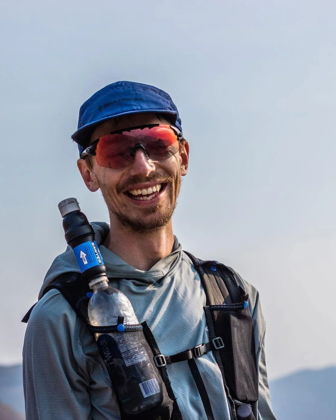 Smiling man wearing sunglasses, a blue cap, and outdoor gear, with a water bottle attached to his backpack.