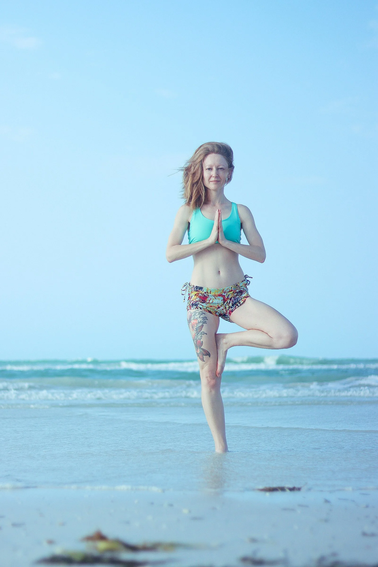 A woman practicing yoga on a beach, standing in tree pose with her hands in prayer position, wearing a turquoise sports top and colorful shorts, with the ocean and sky in the background.