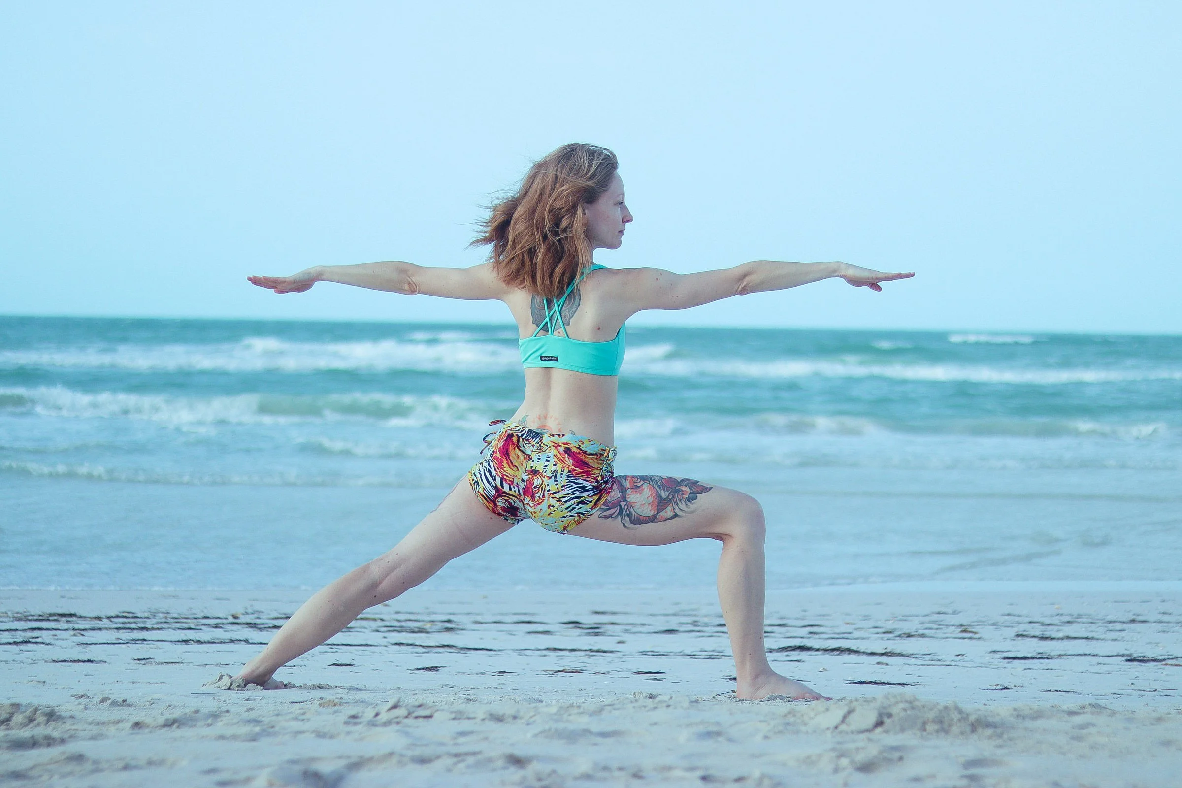 A woman practicing yoga on the beach, standing in a warrior pose with arms extended and one leg bent. She is wearing a turquoise sports bra and colorful shorts, with a tattoo on her thigh, facing the ocean.