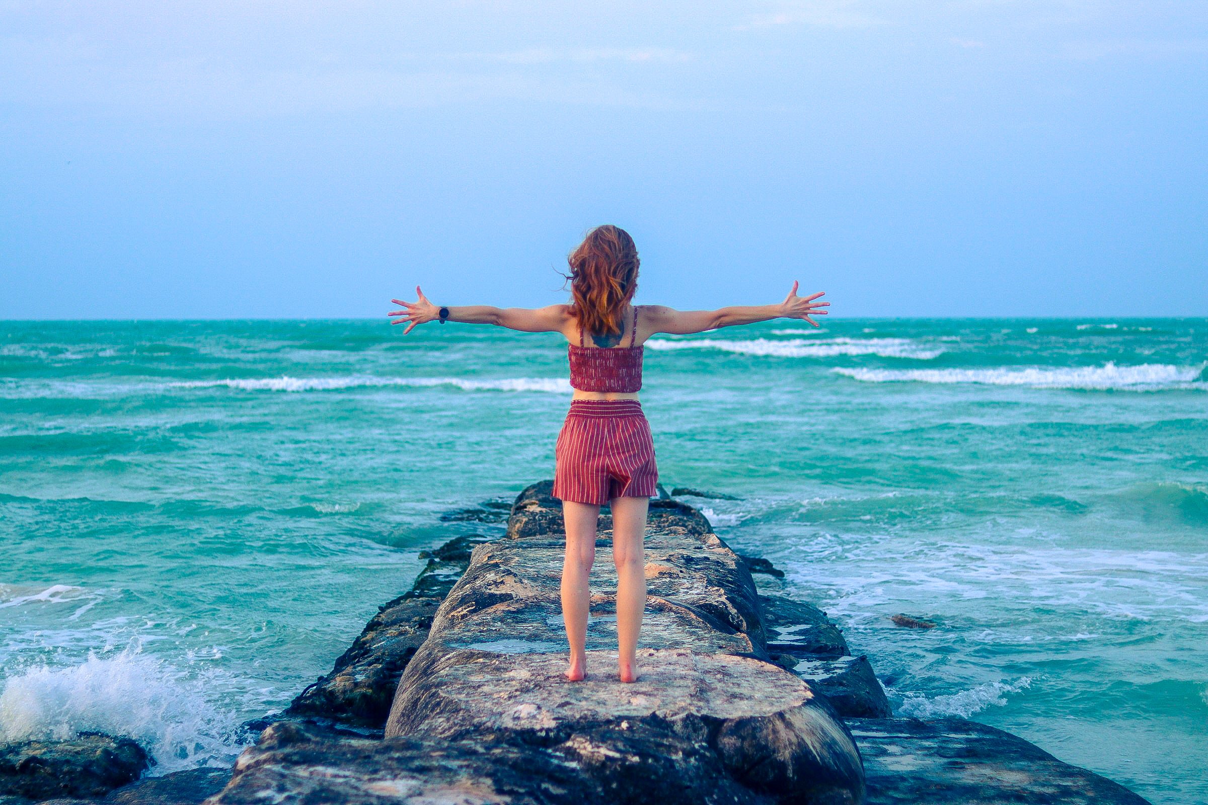 A woman standing on a rocky pier with arms outstretched facing the ocean, enjoying the seaside view.