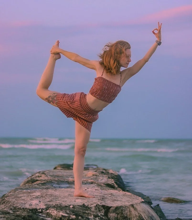 A woman practicing yoga on a rocky pier at the beach during sunset, balancing on one leg in a pose with her other leg bent behind her and her arm reaching forward.