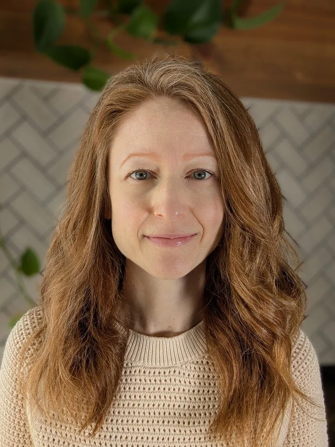 Close-up of a woman with red hair, blue eyes, and a friendly smile, wearing a beige knit sweater, standing indoors with blurred background and some green plants.