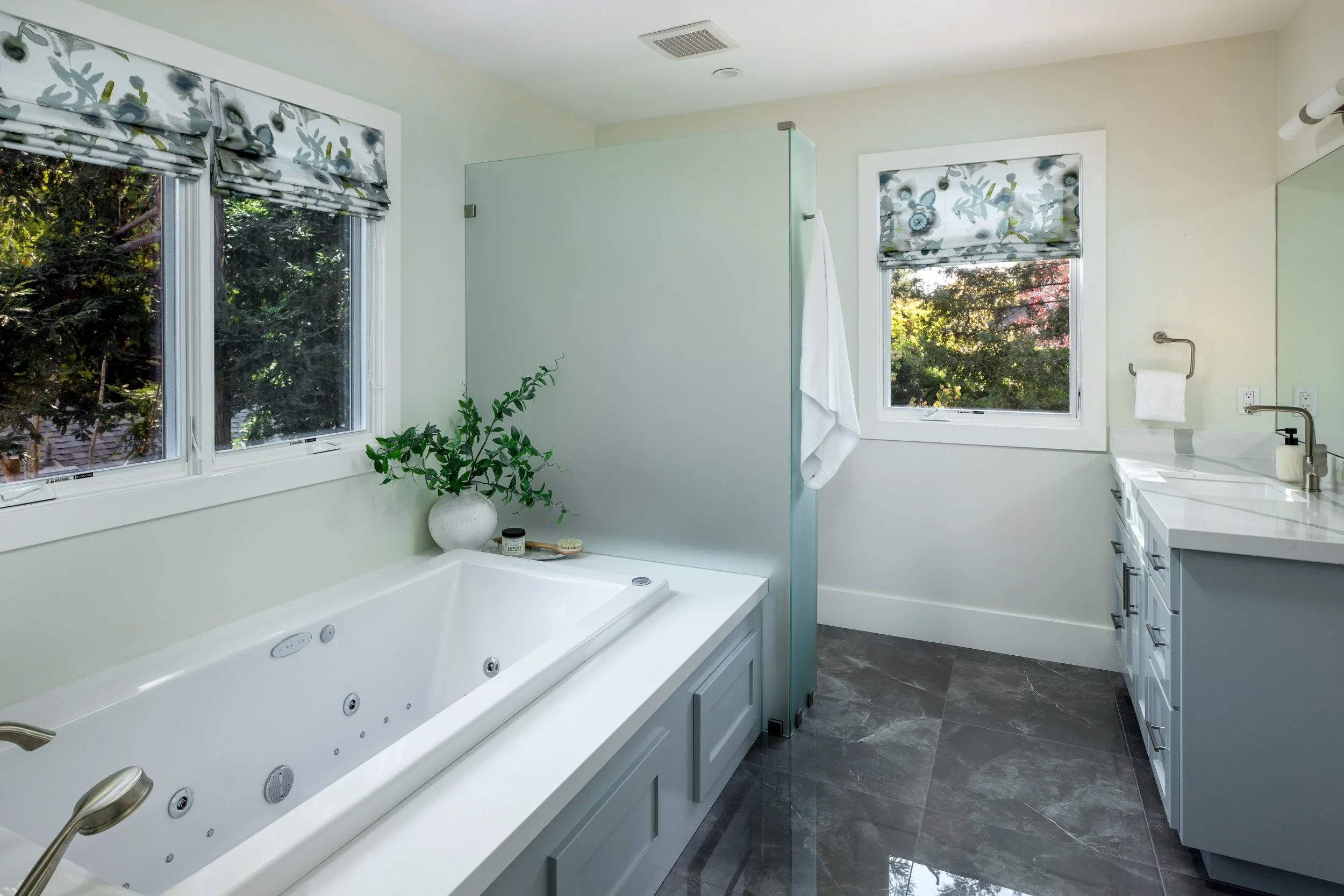 Bathroom with a large bathtub, two windows with floral blinds, a plant in a white vase, a shower enclosure, a white vanity with a sink, and tiled flooring.