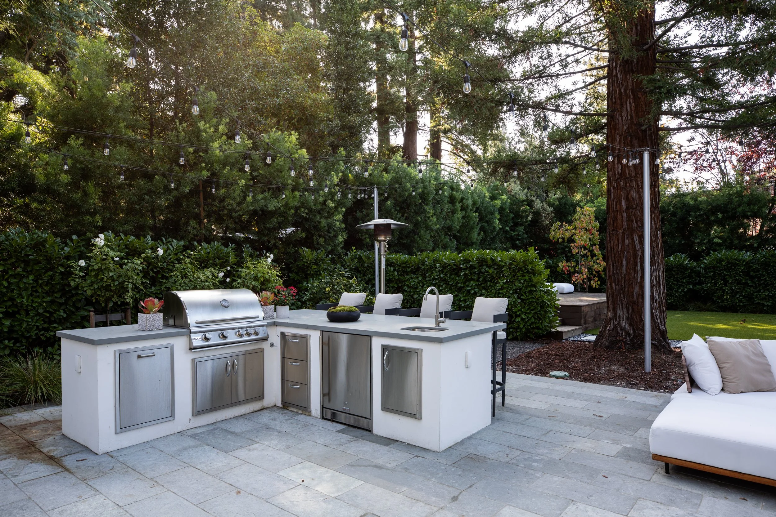 Outdoor kitchen with a built-in grill, countertop, and backyard seating area surrounded by tall trees and greenery, with string lights hanging above.