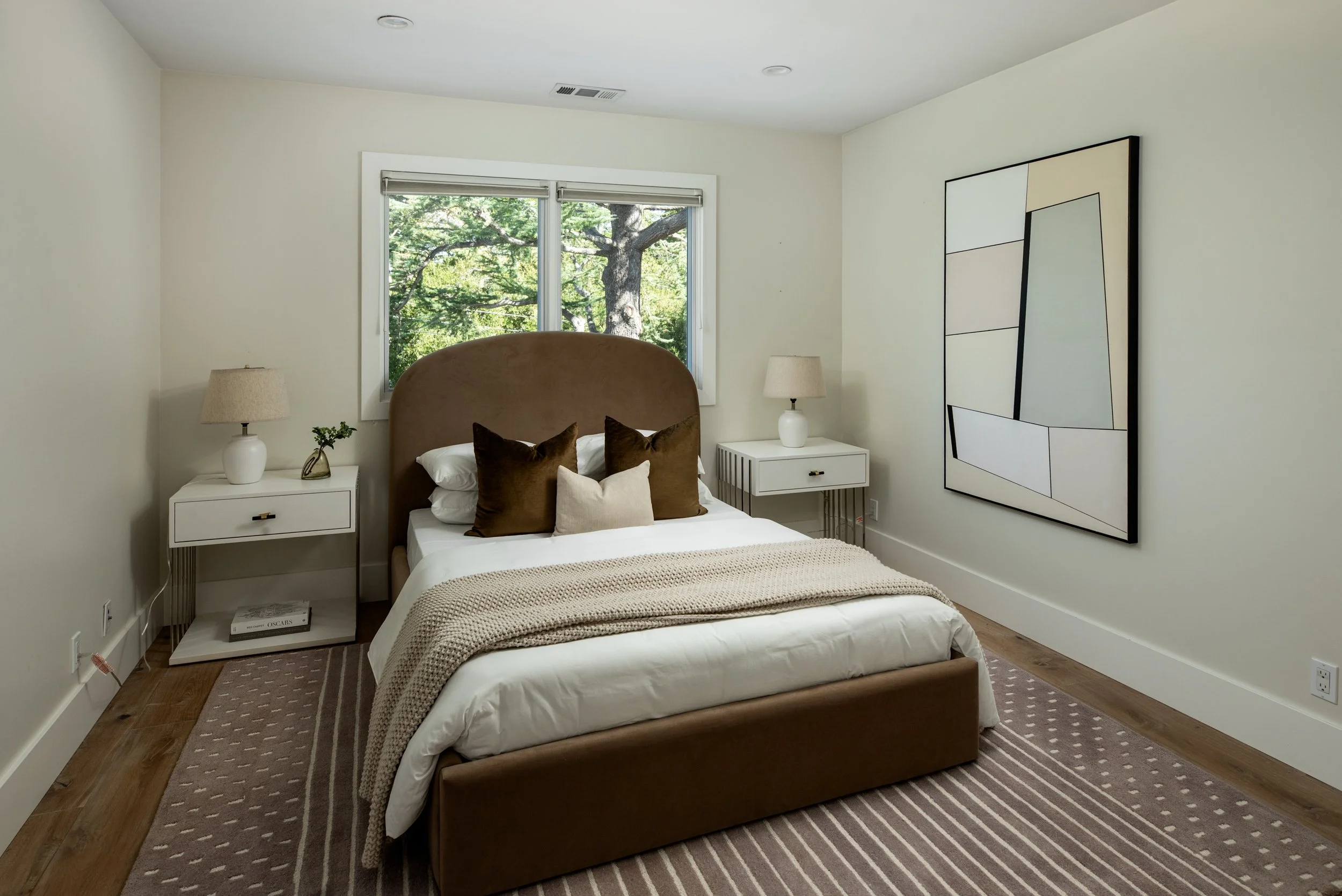 A bedroom with a bed, white bedding, brown and cream pillows, beige blanket, white nightstands with lamps, a small plant, and modern artwork on the wall; window with glimpses of green trees outside.