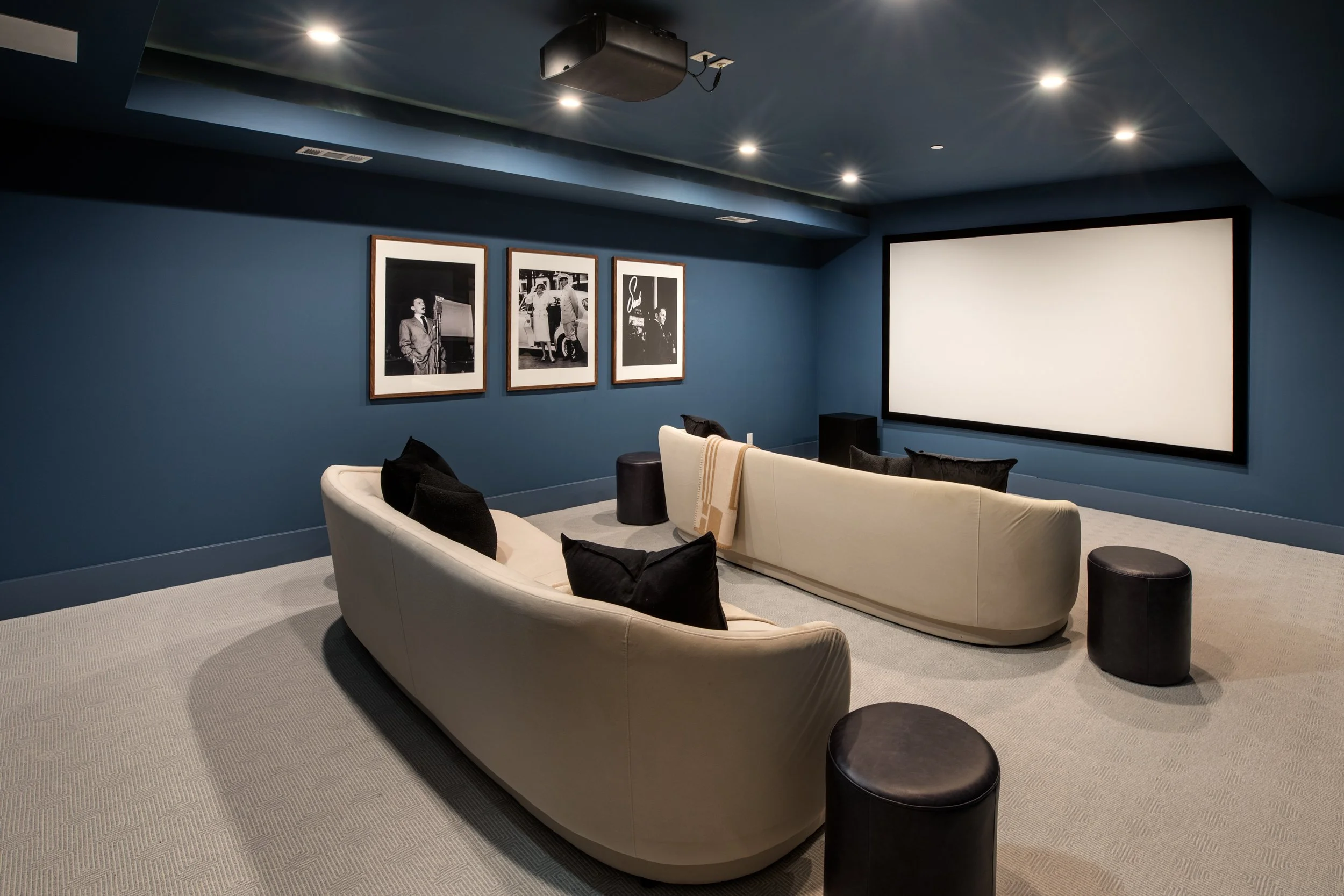 A modern home theater room with a large white sofa, black pillows, a framed black-and-white photos on the blue wall, a projector mounted on the ceiling, and a large blank screen on the wall.