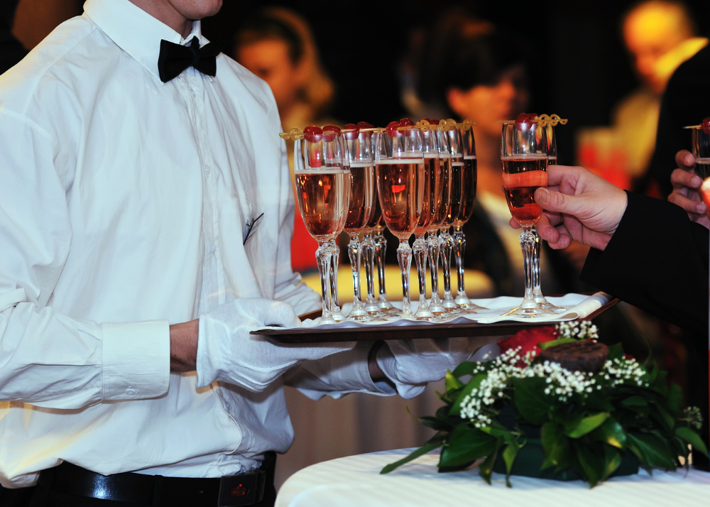 A server in a white shirt, black bow tie, and white gloves is holding a tray with multiple glasses of pink champagne, which is being served to guests at a formal event.