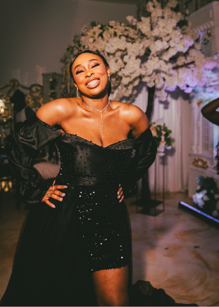 A woman in a black sequined dress with off-the-shoulder puffed sleeves smiling at a formal event, with a background of white flowers and ornate decorations.