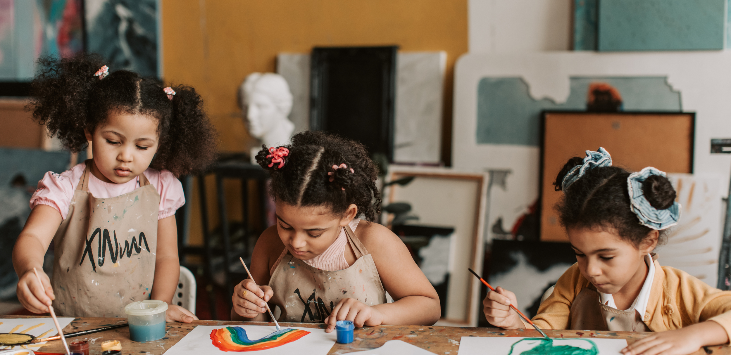Three young girls painting rainbows on paper at an art studio, wearing aprons, with art supplies and paintings in the background.