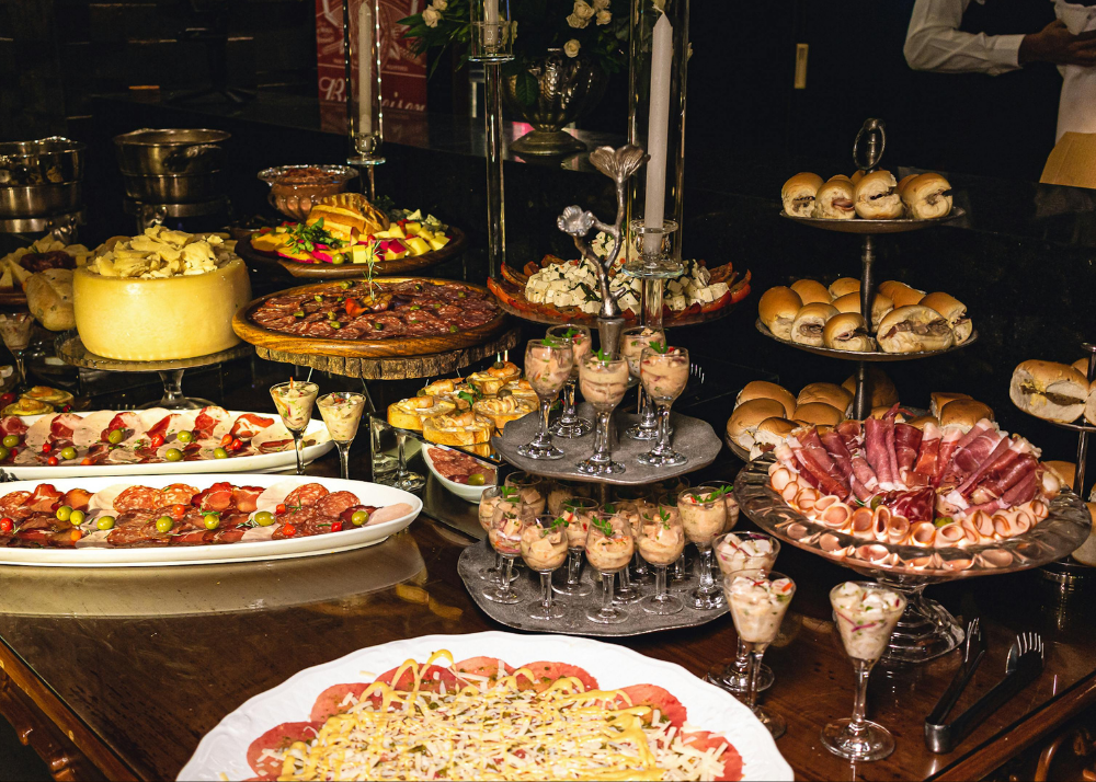 A variety of appetizers and finger foods displayed on a dark table, including mini sandwiches, cold cuts, cheese, fruit, and desserts.