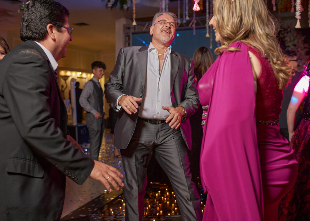 People dancing and enjoying themselves at a lively celebration or party, with one man in a gray suit laughing and dancing with women in pink and maroon dresses.