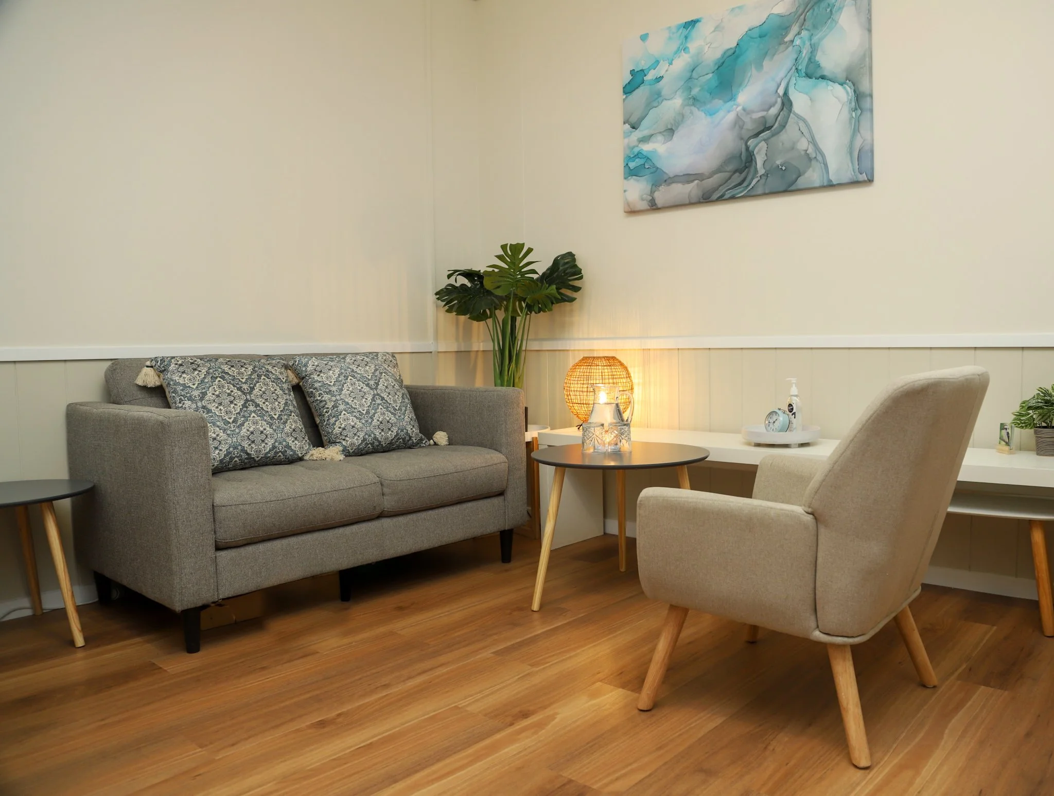 A cozy living room corner with a gray sofa, patterned throw pillows, a beige armchair, a round coffee table with decorative candles, a wicker table lamp, a white shelf with a small clock and plants, and a blue abstract painting on the wall.