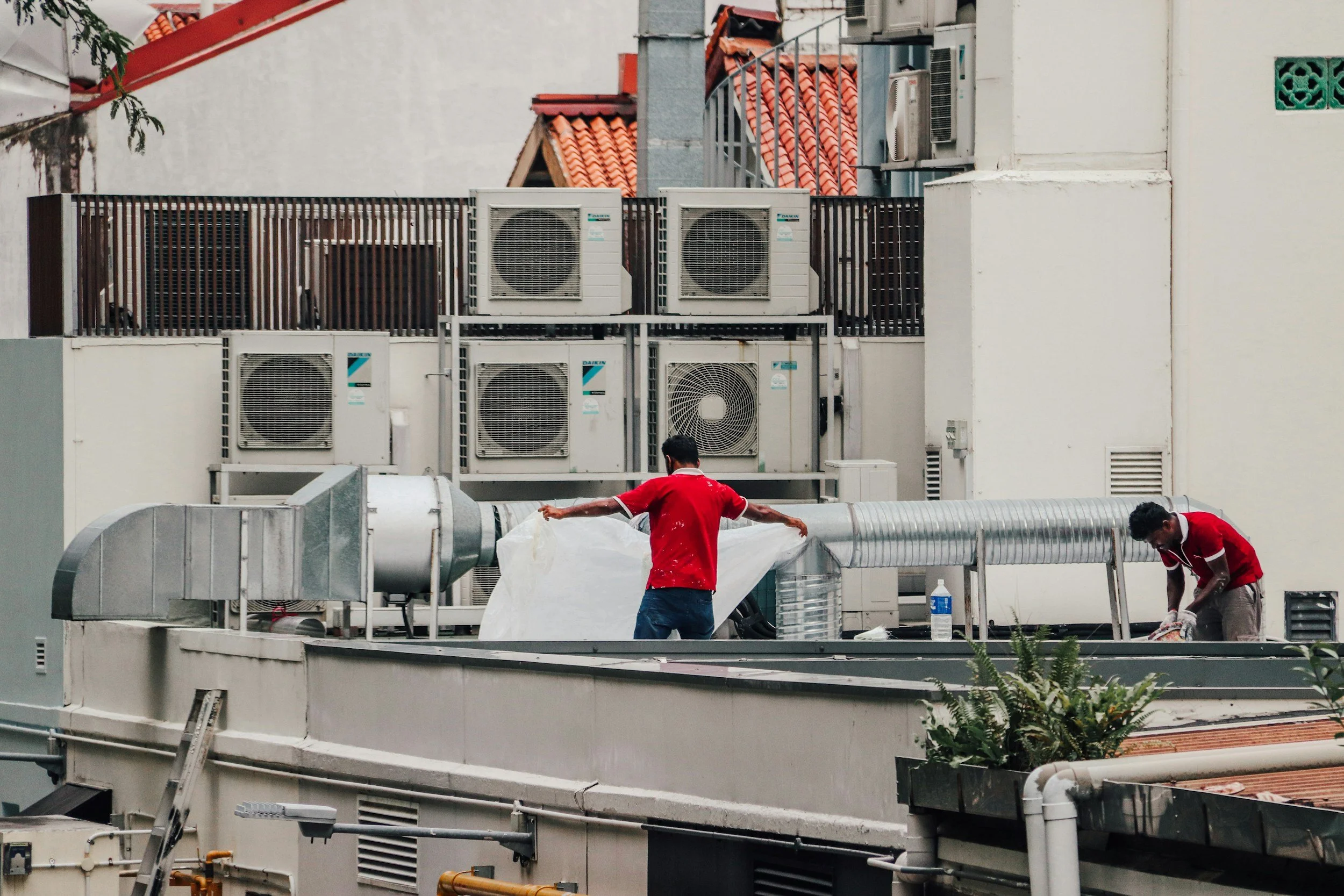 Two workers on a rooftop, wearing red shirts, are installing or repairing ventilation ductwork. The rooftop has several air conditioning units and a water bottle is visible.