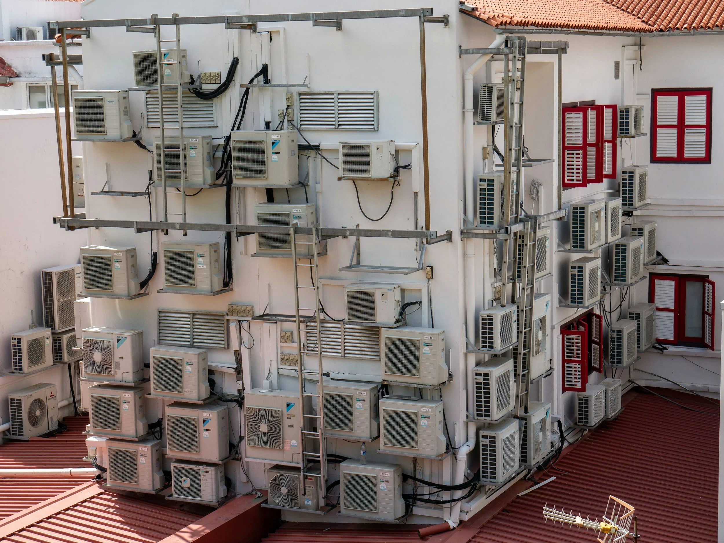 A building exterior with multiple air conditioning units installed on the wall and scaffolding supporting some units, alongside red window frames with white shutters.