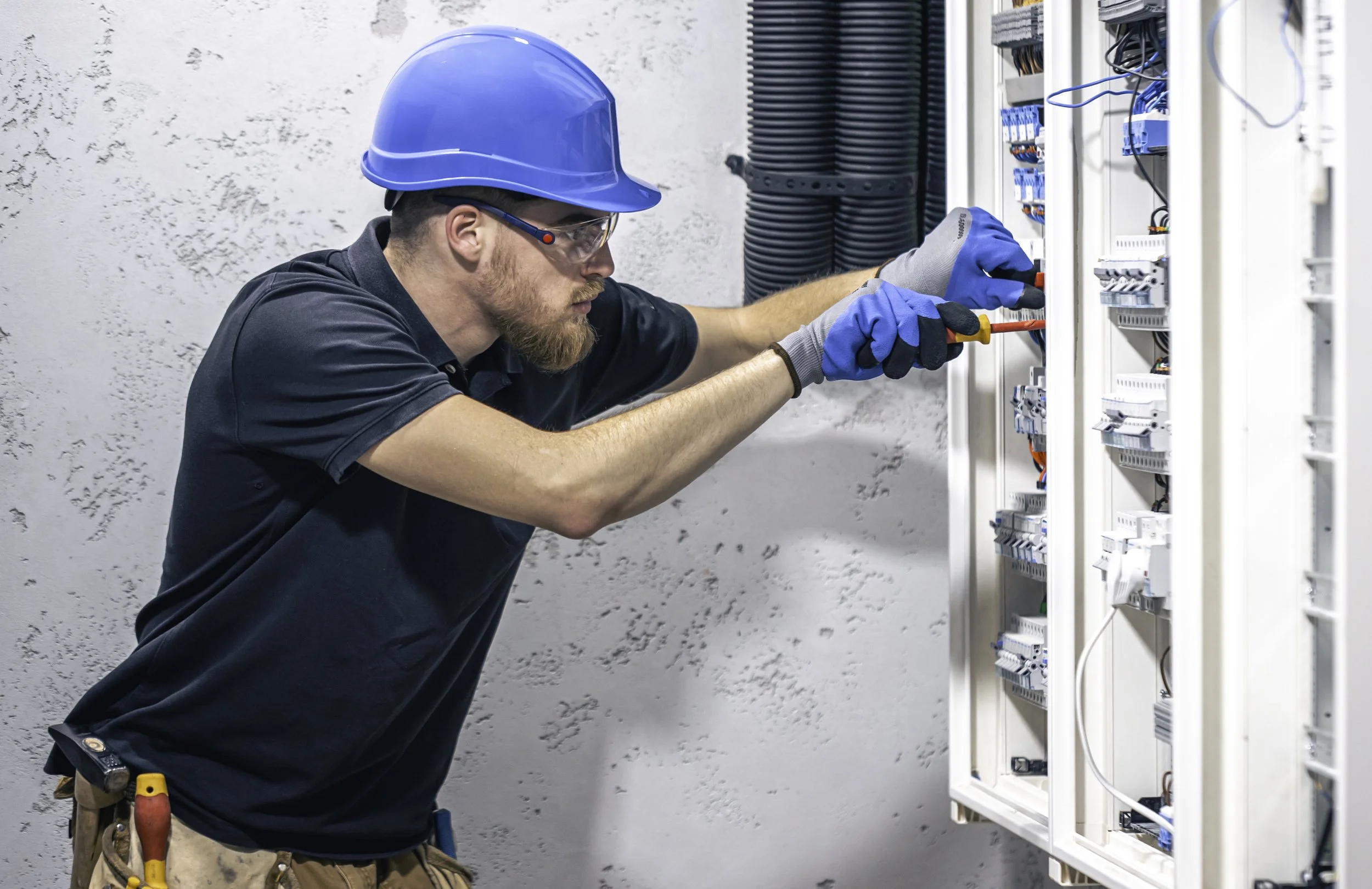 A male electrician wearing a blue helmet, safety glasses, gloves, and a black shirt working on an electrical panel with tools in his belt.