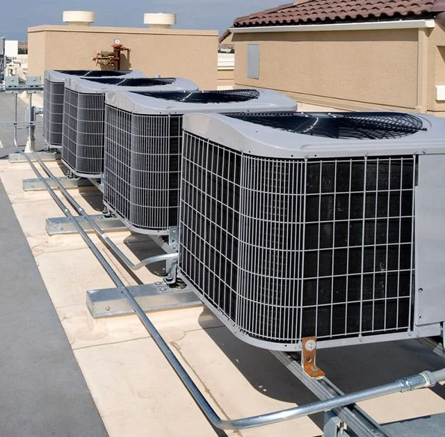 Four large commercial HVAC units on a flat rooftop with ductwork and piping, against beige building walls and a roof with reddish shingles.