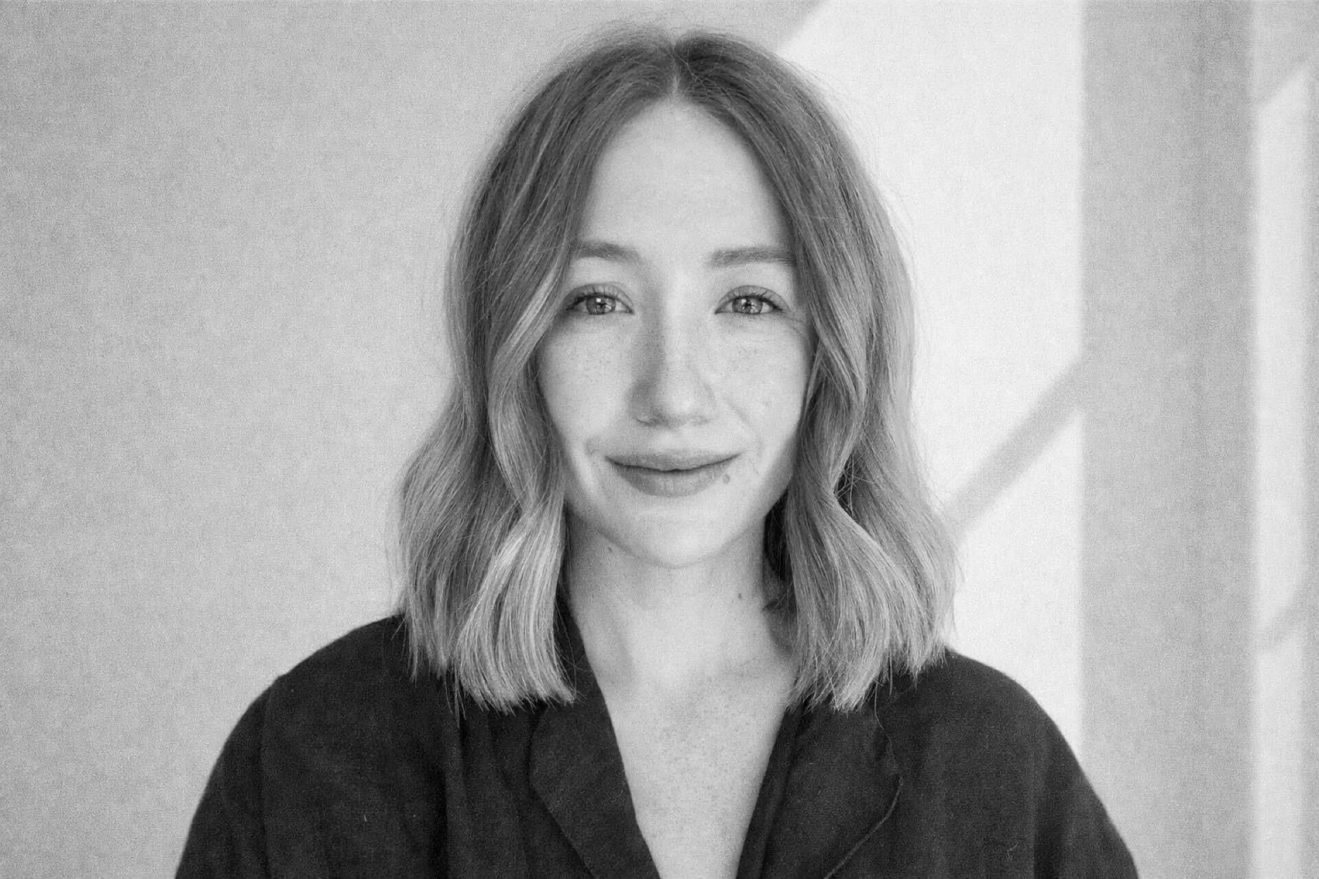 Black and white portrait of Emma, a young woman with shoulder-length wavy hair, smiling softly, wearing a dark shirt, in front of a plain background.