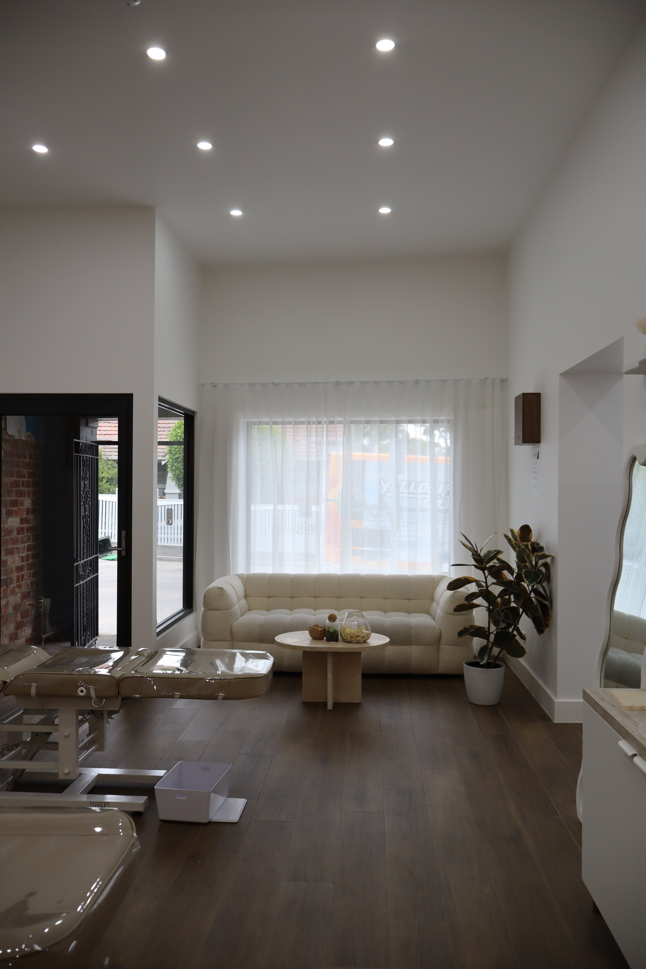 A waiting room or lounge with a cream-colored tufted sofa, a small round wooden coffee table with decorative items, a large potted plant, a mirror on the wall, and overhead recessed lighting. There's a window with sheer curtains allowing natural ligh