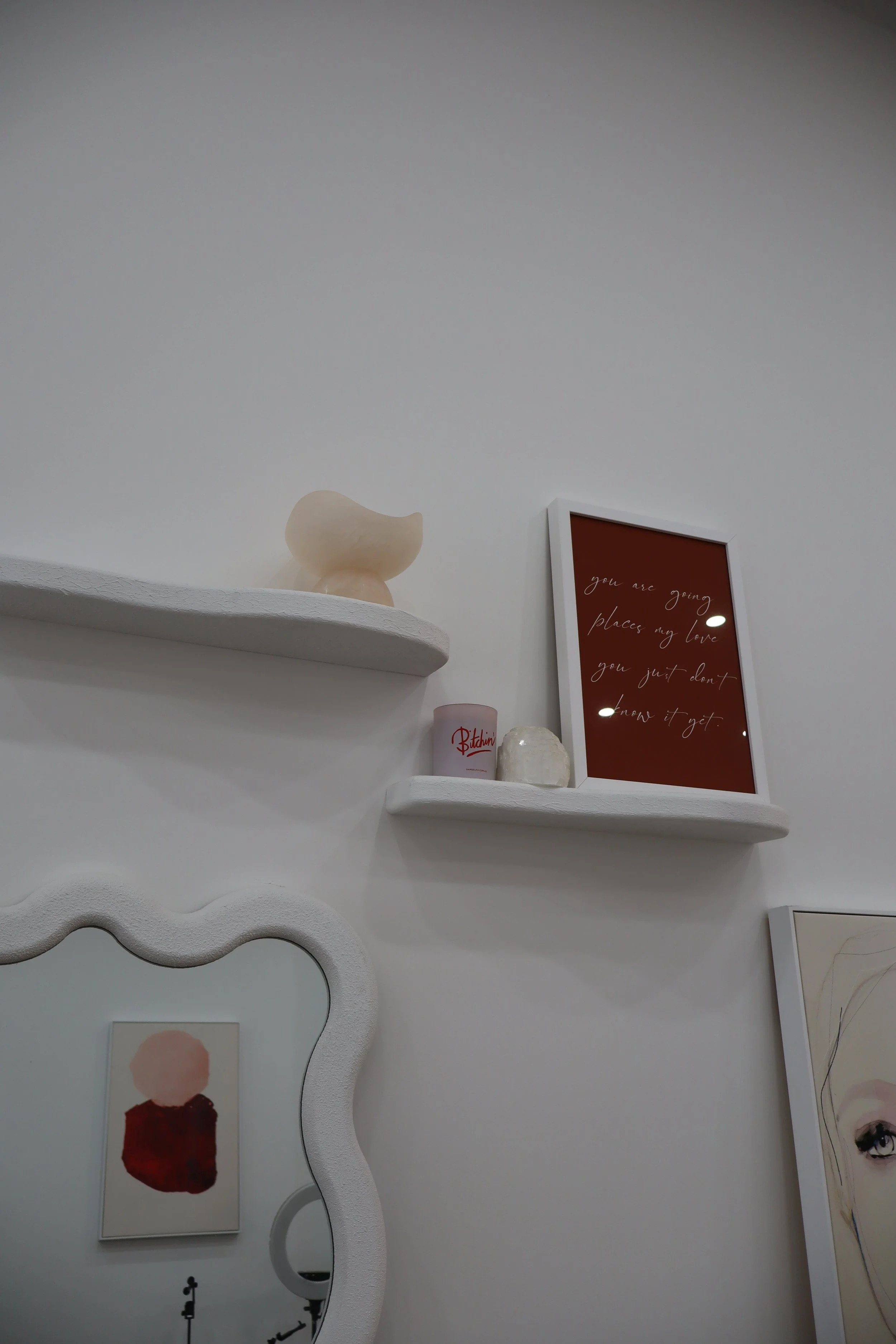 Decorative wall shelves with a ceramic vessel, a framed quote, a candle, and a stone, along with artwork and a mirror in a minimalist aesthetic.