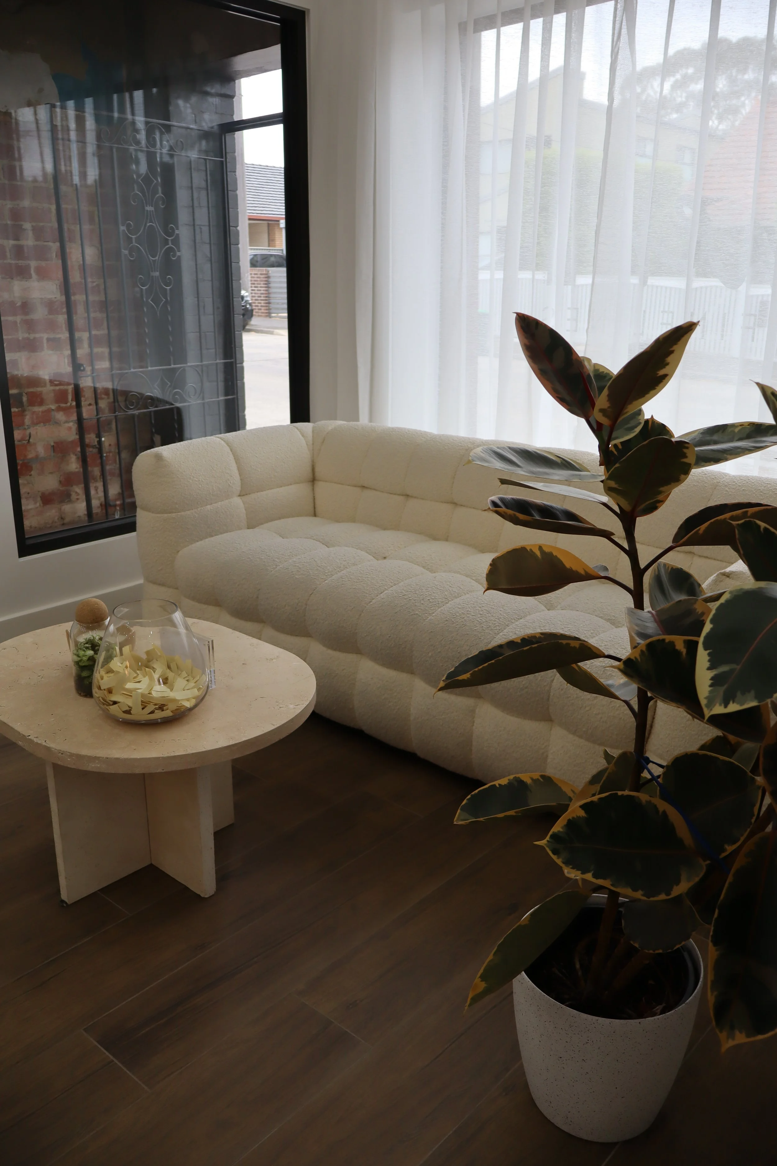 A cozy sitting area with a beige tufted sofa, a small round coffee table with decorative items, a large leafy plant in a white pot, and sheer curtains letting in natural light.