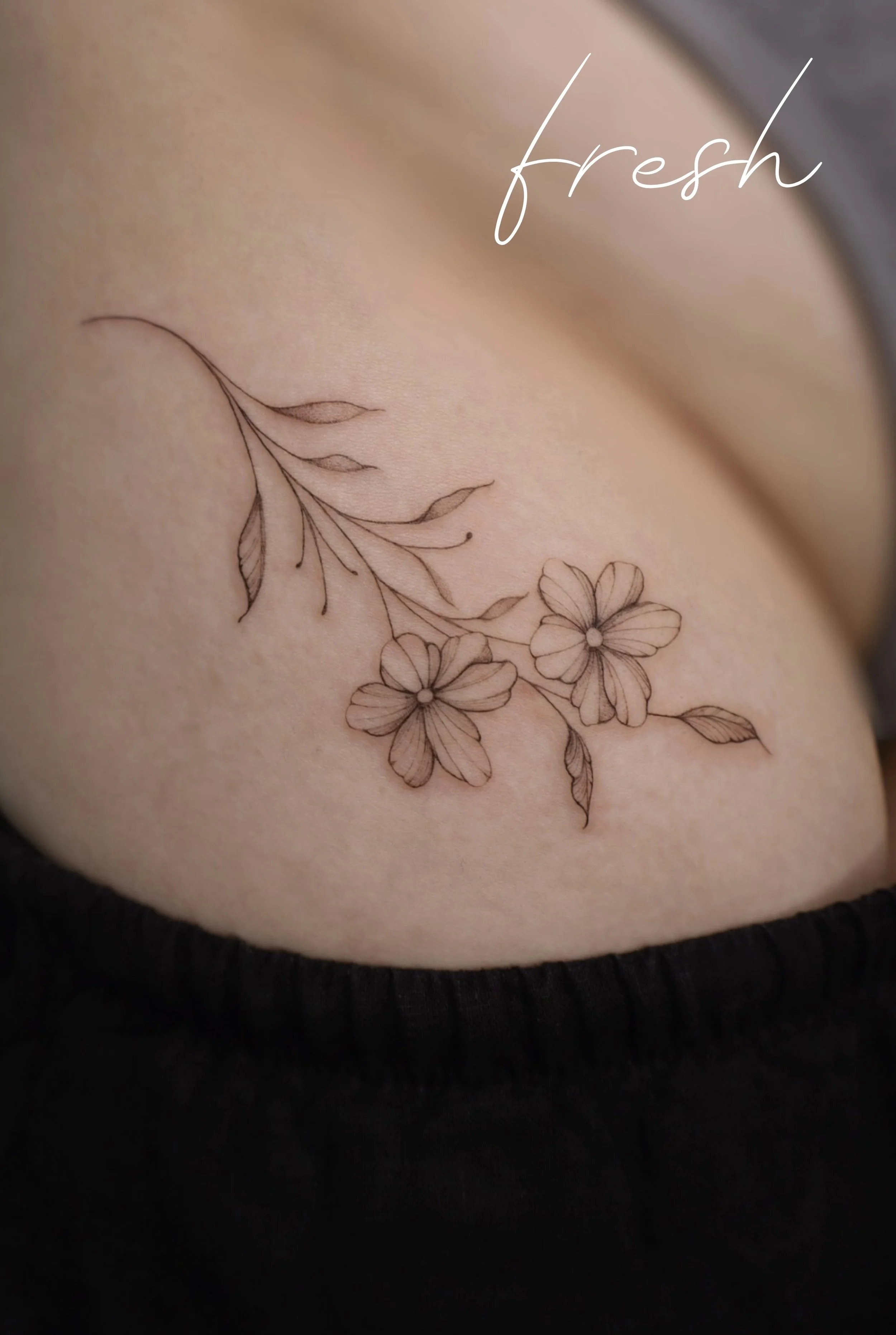 A tattoo of a delicate floral design with two flowers and leaves on skin, with the word 'fresh' written in cursive.