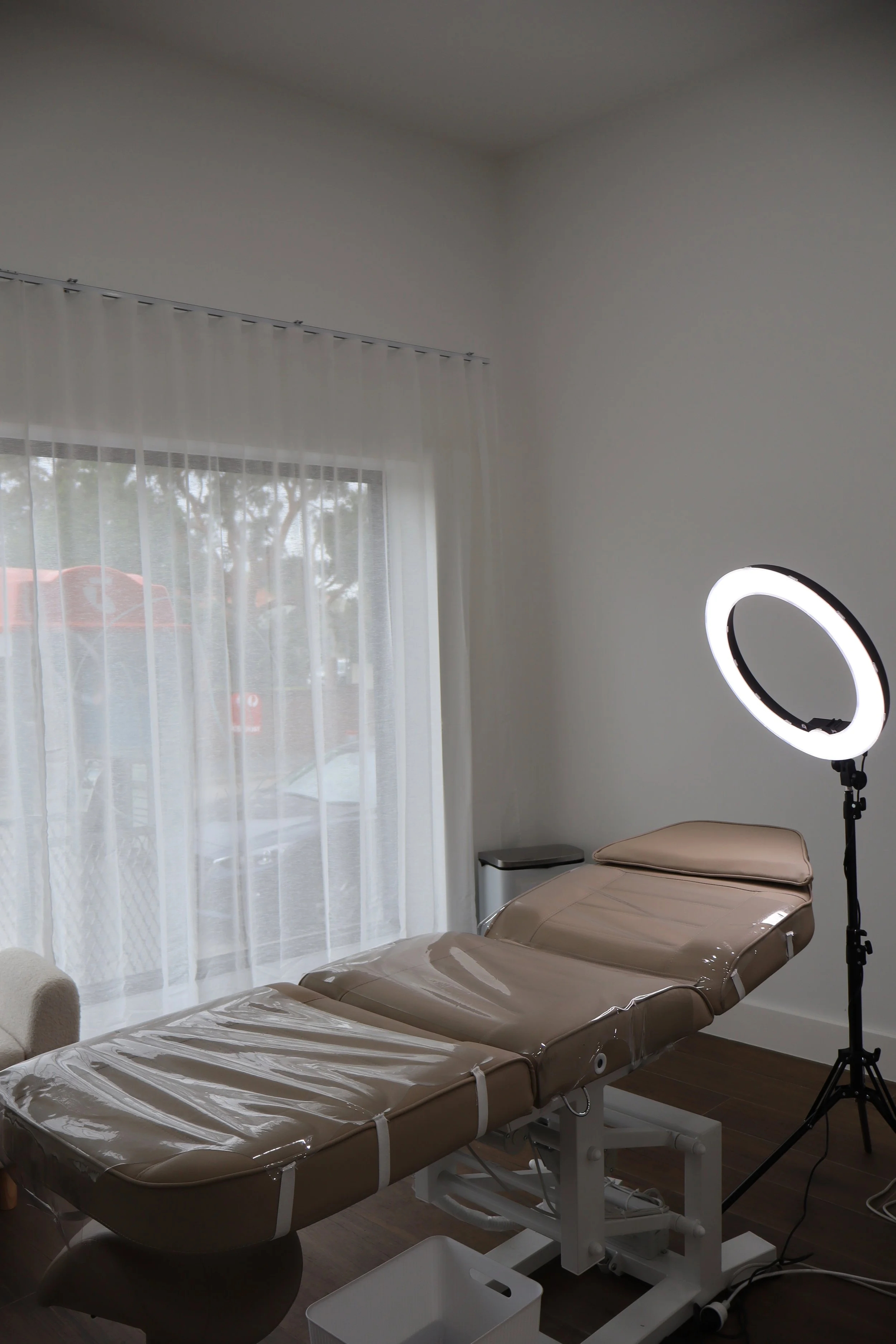 Medical examination room with an adjustable beige examination table, a ring light on a stand, a small trash can, a window with sheer white curtains, and hardwood flooring.