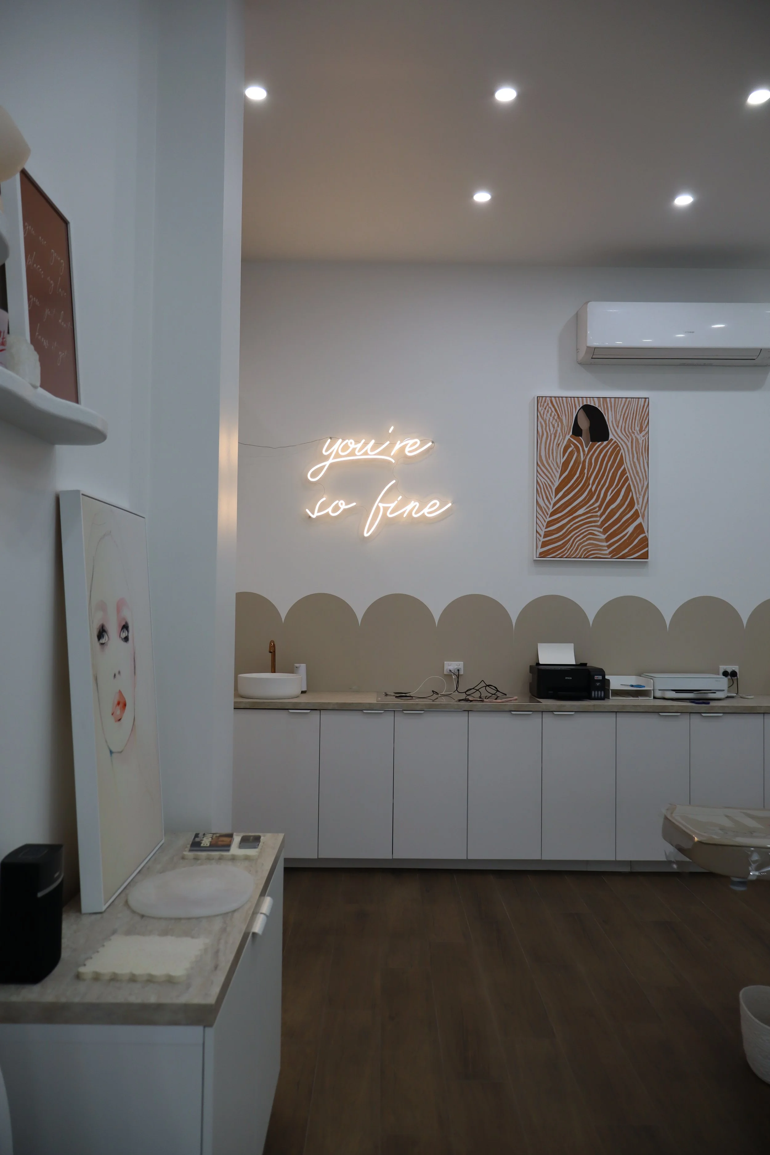 Interior of a modern room with white walls, a neon sign that reads "you're so fine," a painting of a woman with black hair and a patterned orange dress, a white countertop with a small white sink, and various electronic devices including a printer an