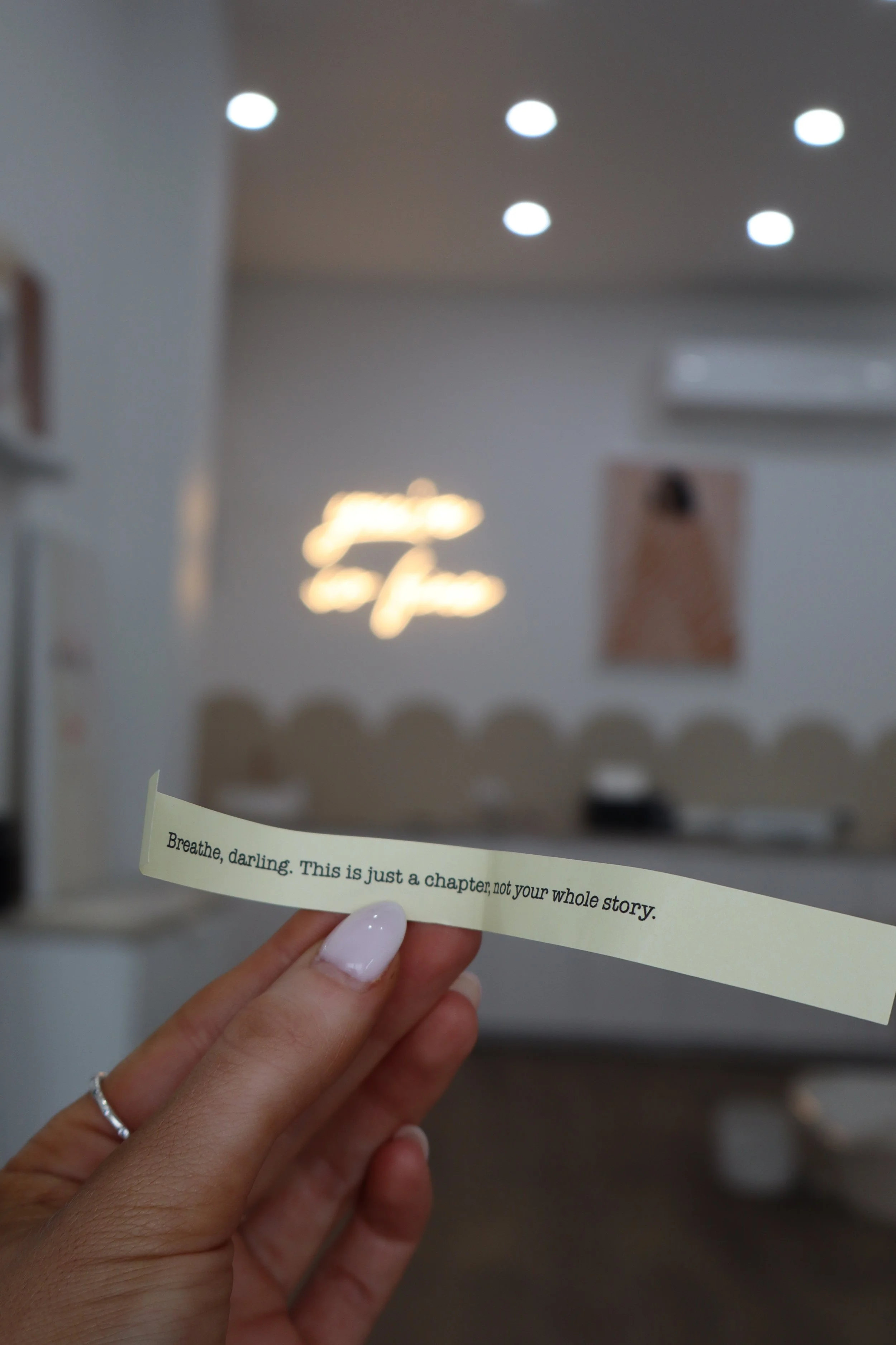 A person holding a small printed quote note that reads, 'Breathe, darling. This is just a chapter, not your whole story,' inside a modern, softly lit room with blurred background.