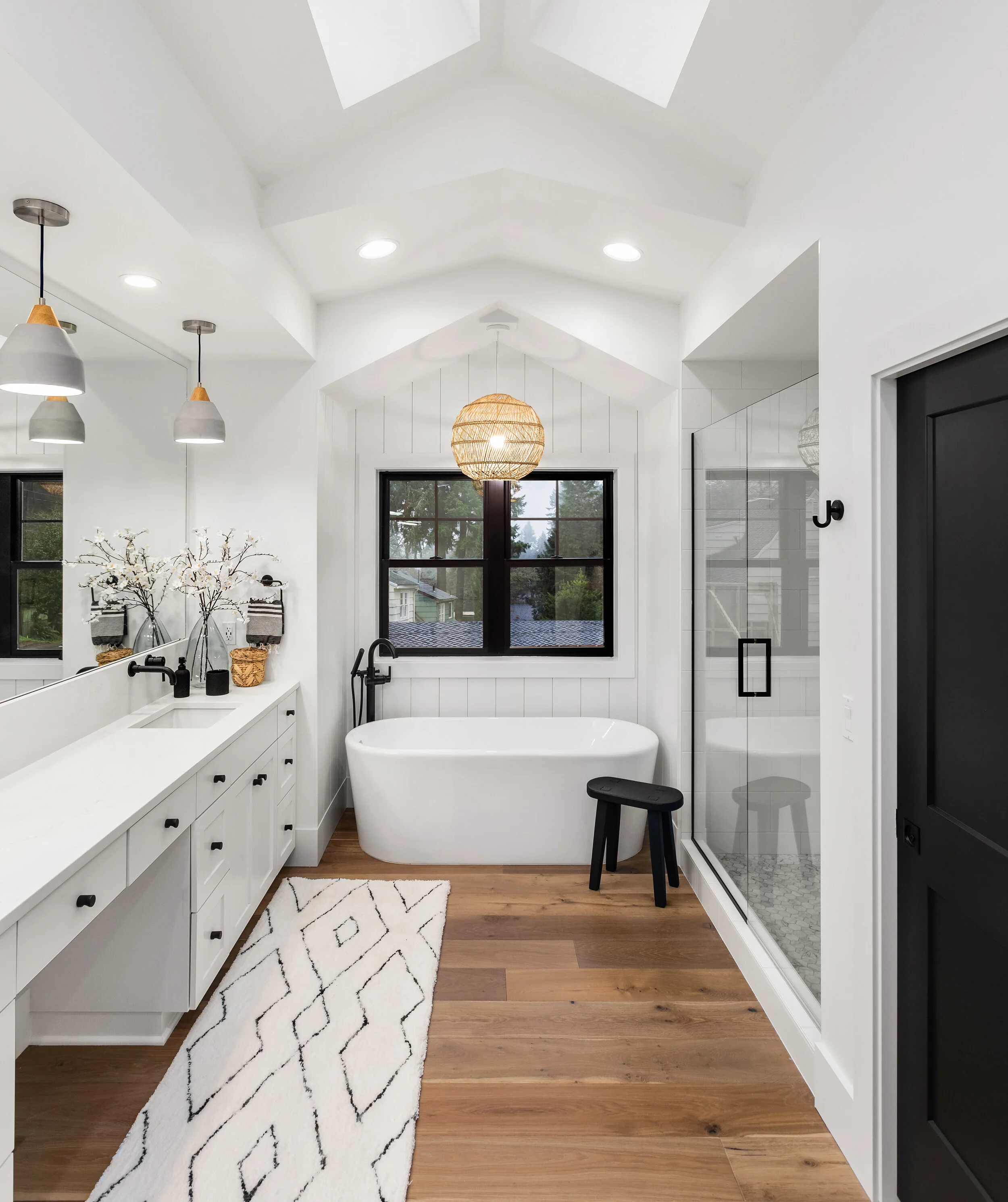 Modern bathroom with white bathtub, glass-enclosed shower, long vanity with sink, decorative mirror, black fixtures, wooden floor, patterned rug, and pendant lighting.