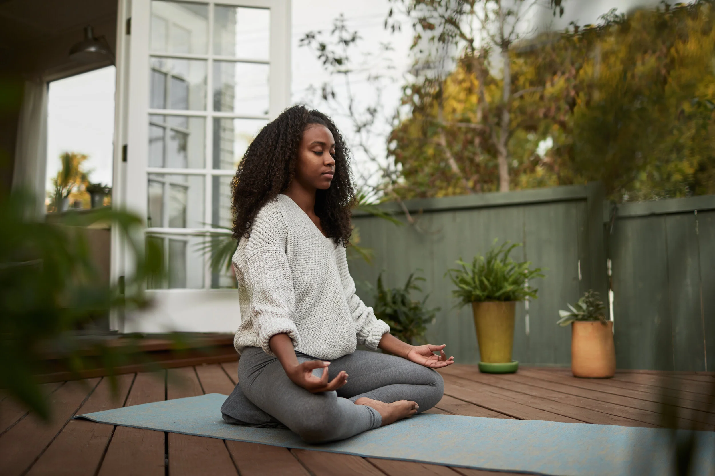 Woman practicing yoga meditation on a mat outdoors on a wooden deck, wearing a gray sweater and leggings, with potted plants nearby.