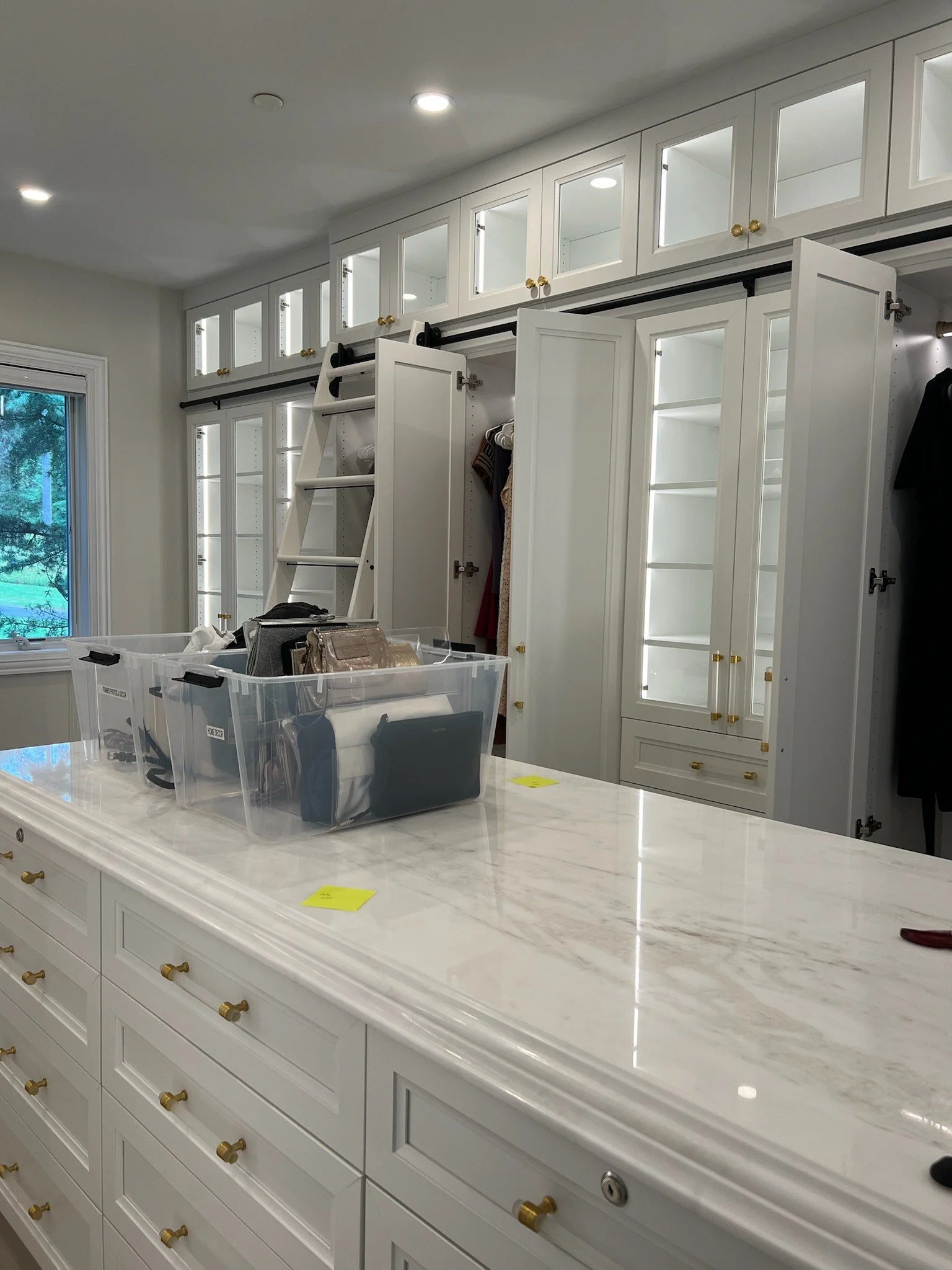 Luxury walk-in closet with white cabinets and marble island, featuring storage bins, hanging clothes, and a ladder for upper cabinets. Professionally organized by Uncluttered Spaces located in Bucks County. 