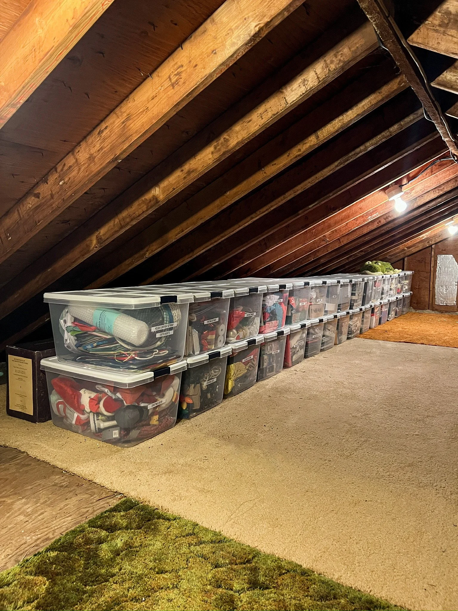 Attic with wooden beams and organized plastic storage bins lined up along the wall, containing assorted holiday decorations. Professionally organized by Uncluttered Spaces located in Bucks County. 