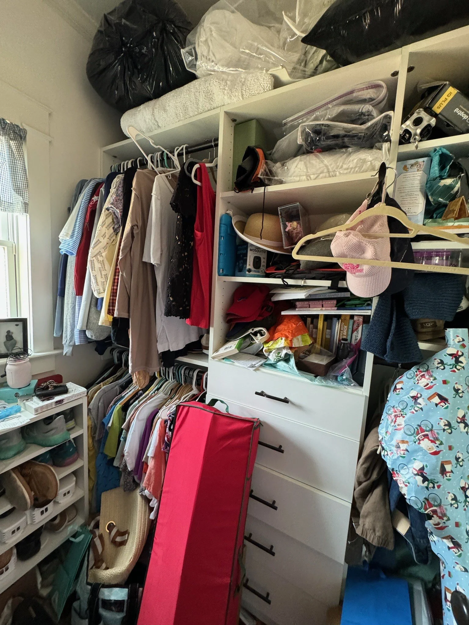 A cluttered closet with clothes hanging, shelves filled with bags, hats, and miscellaneous items. Shoes are organized on racks and various storage containers are visible. A red bag leans against a dresser. Professionally organized by Uncluttered Spac