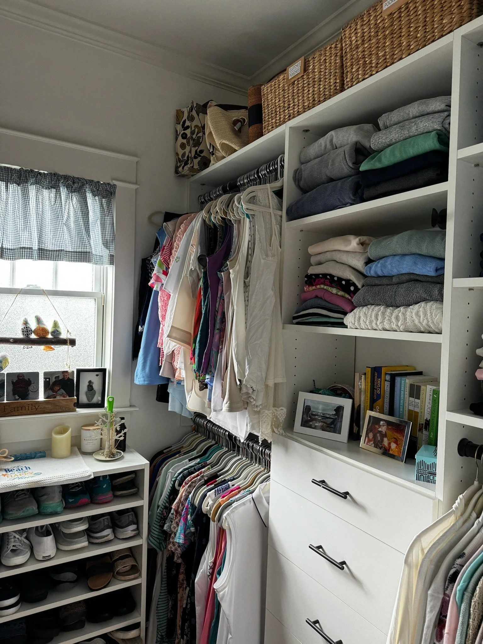 Closet with organized clothing on hangers, folded sweaters on shelves, and shoes on racks. Baskets and books are on upper shelves, and family photos are displayed near a window with a checkered curtain. Professionally organized by Uncluttered Spaces 
