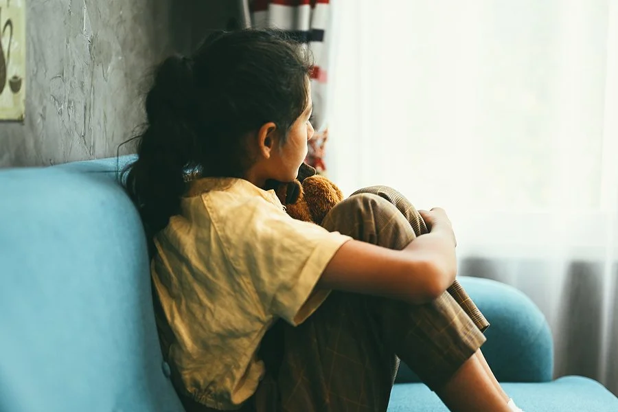 Child sitting on a blue couch hugging a stuffed toy, looking out the window.
